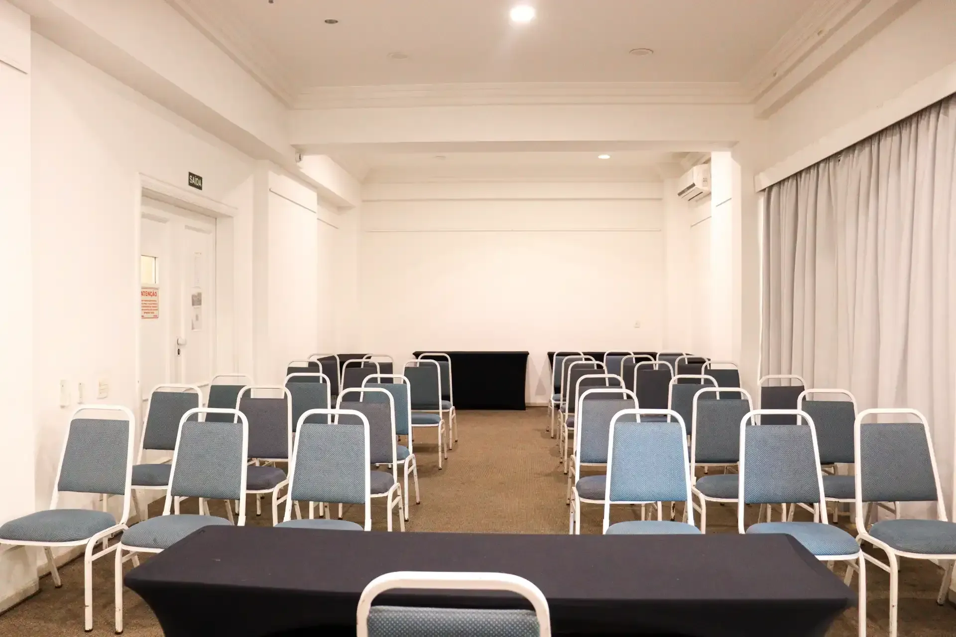 Sala de conferências vazia com fileiras de cadeiras azuis voltadas para uma mesa e paredes brancas.