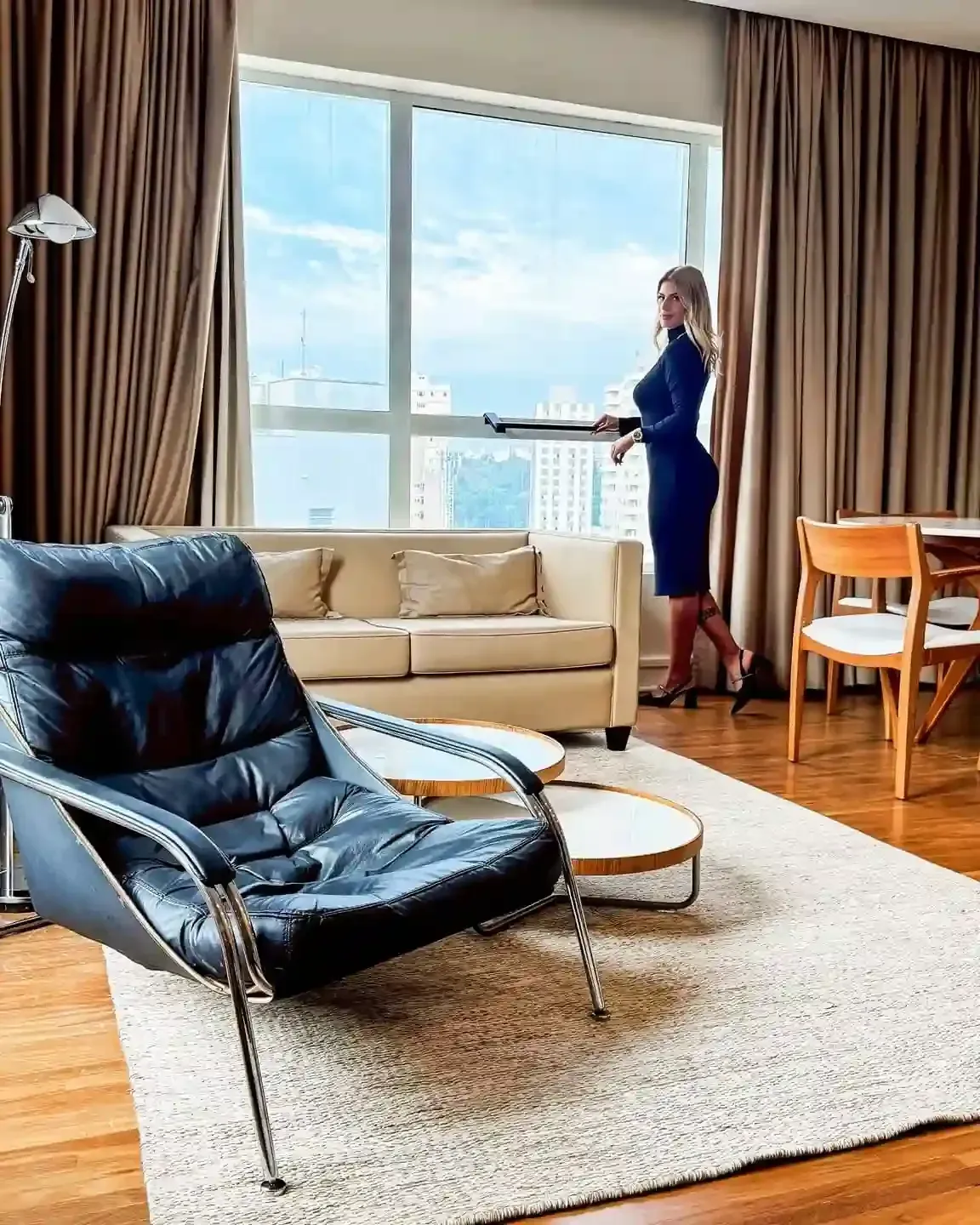 Mulher de vestido azul olha pela janela. Sala de estar com mobília moderna, vista para a cidade.