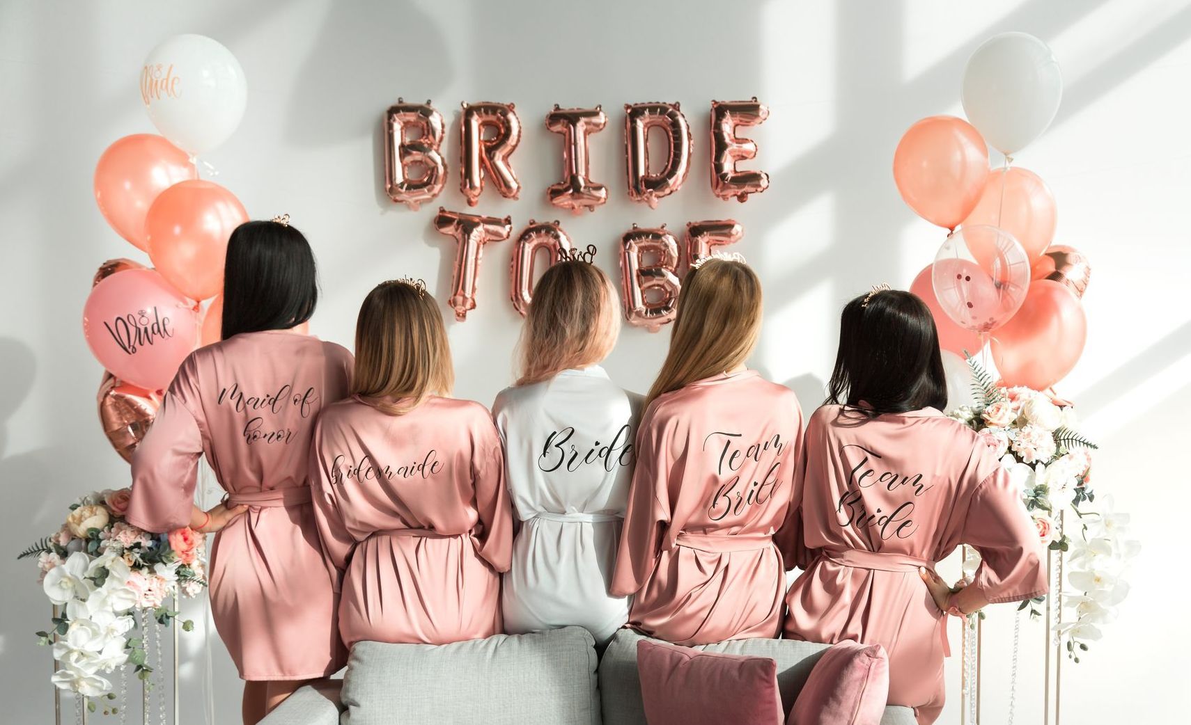 Cinco mulheres de robes rosa estão de costas em uma festa de despedida de solteira, de frente para balões dourados com a inscrição
