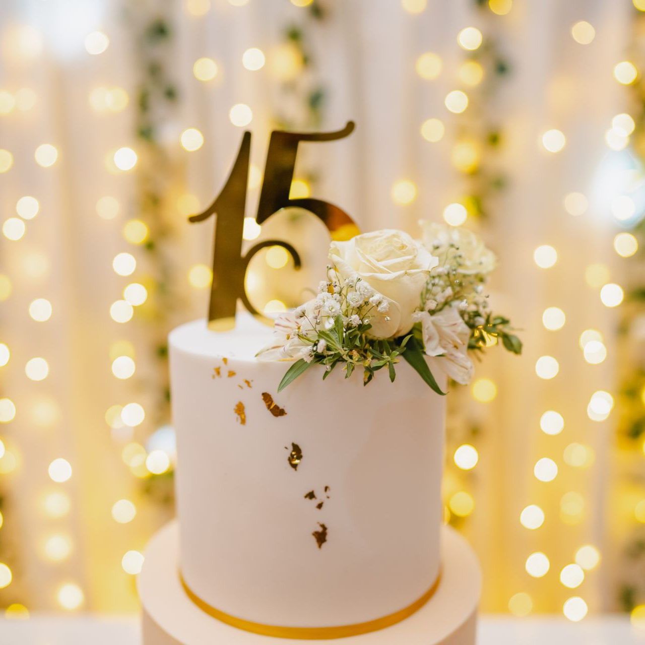 Bolo branco de 15º aniversário com topo dourado e flores, sob luzes bokeh em tons quentes.