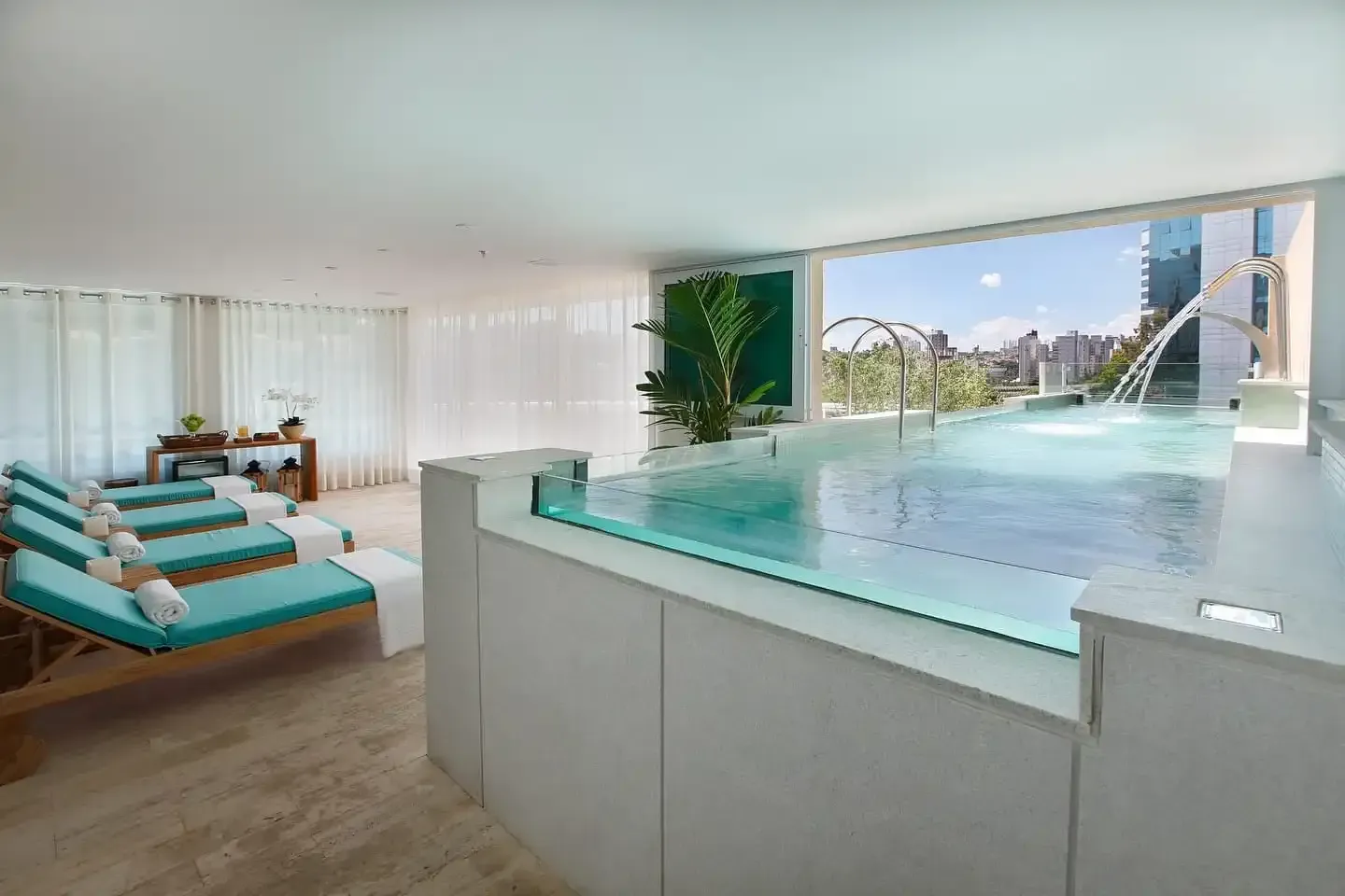 Área de piscina coberta com espreguiçadeiras turquesa, piscina com laterais de vidro e vista para a cidade.