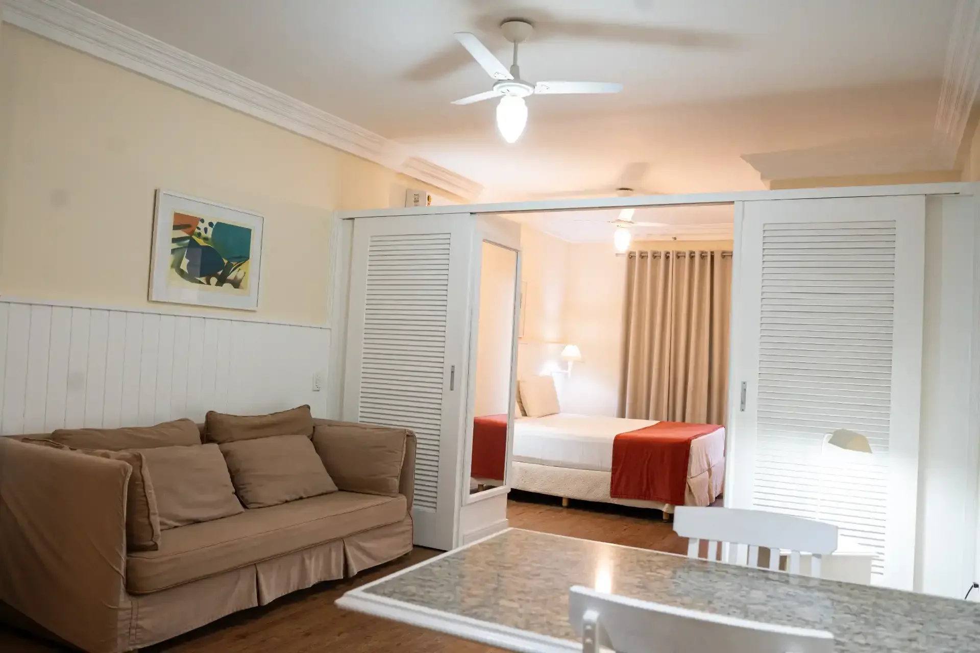 Sala de estar iluminada com sofá bege, persianas brancas e um quarto visível através de portas abertas.