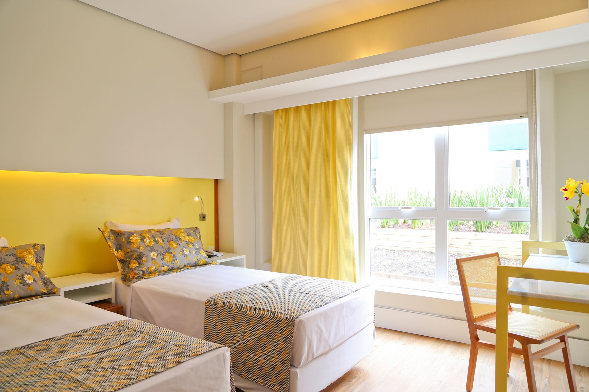 Quarto de hotel luminoso com duas camas de solteiro, detalhes em amarelo e uma área de estar junto à janela.