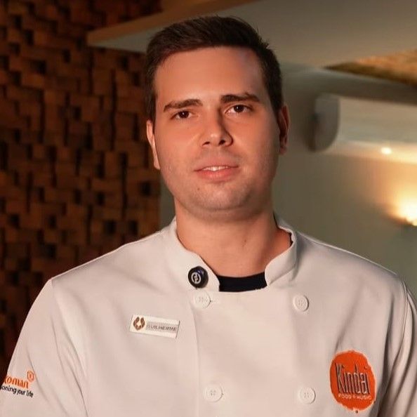 Retrato de um chef de jaqueta branca em pé em um ambiente interno com iluminação quente.