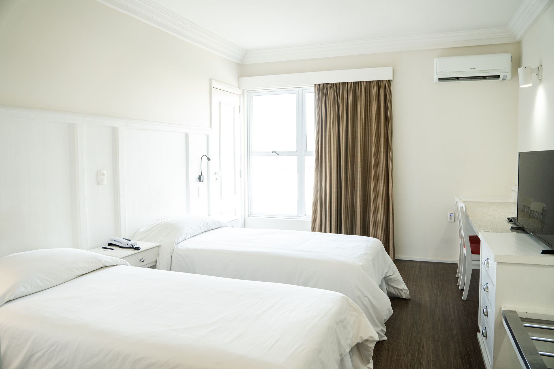 Quarto de hotel luminoso com duas camas brancas, janela com cortinas, TV e ar condicionado.