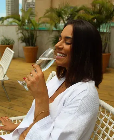 Mulher sorridente de robe branco bebe champanhe sentada em uma cadeira de pátio entre palmeiras em vasos.