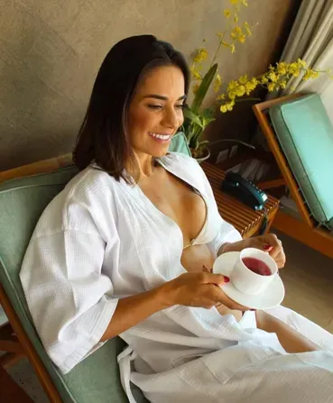 Mulher de robe branco sorrindo enquanto segura uma xícara de chá em um sofá, com flores ao fundo.