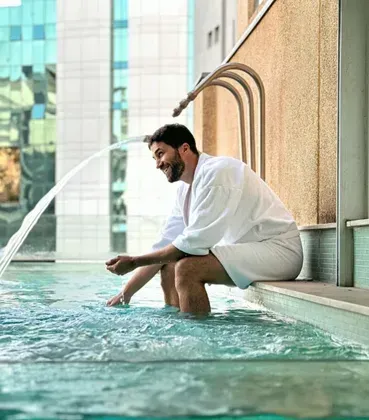 Homem relaxando em uma piscina na cobertura, com os pés na água, ao lado de prédios de vidro e uma parede bege.