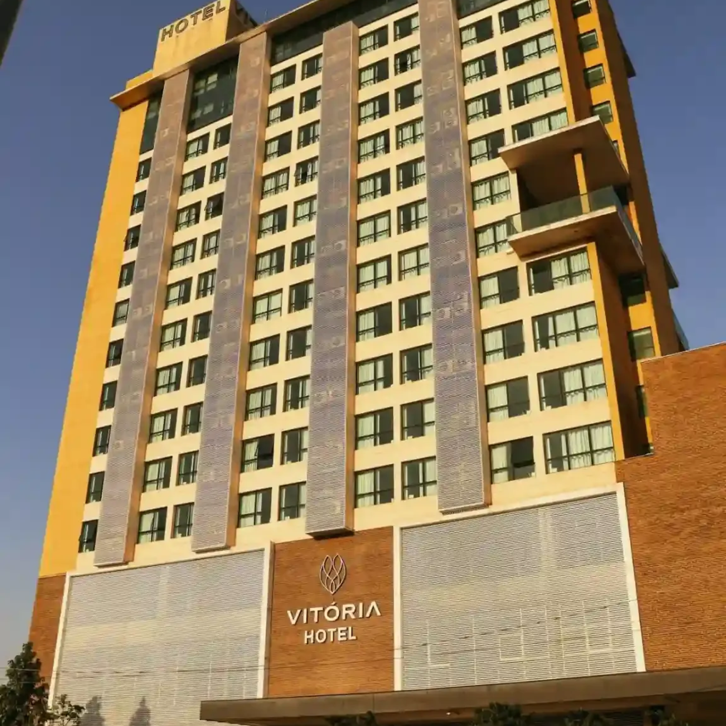 Exterior do Hotel Vitória, um prédio alto em tons de bege e marrom com uma placa bem visível perto da entrada.