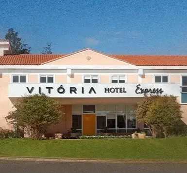 Edifício do Hotel Vitoriá Express com telhado vermelho, fachada branca e gramado verde sob um céu claro.