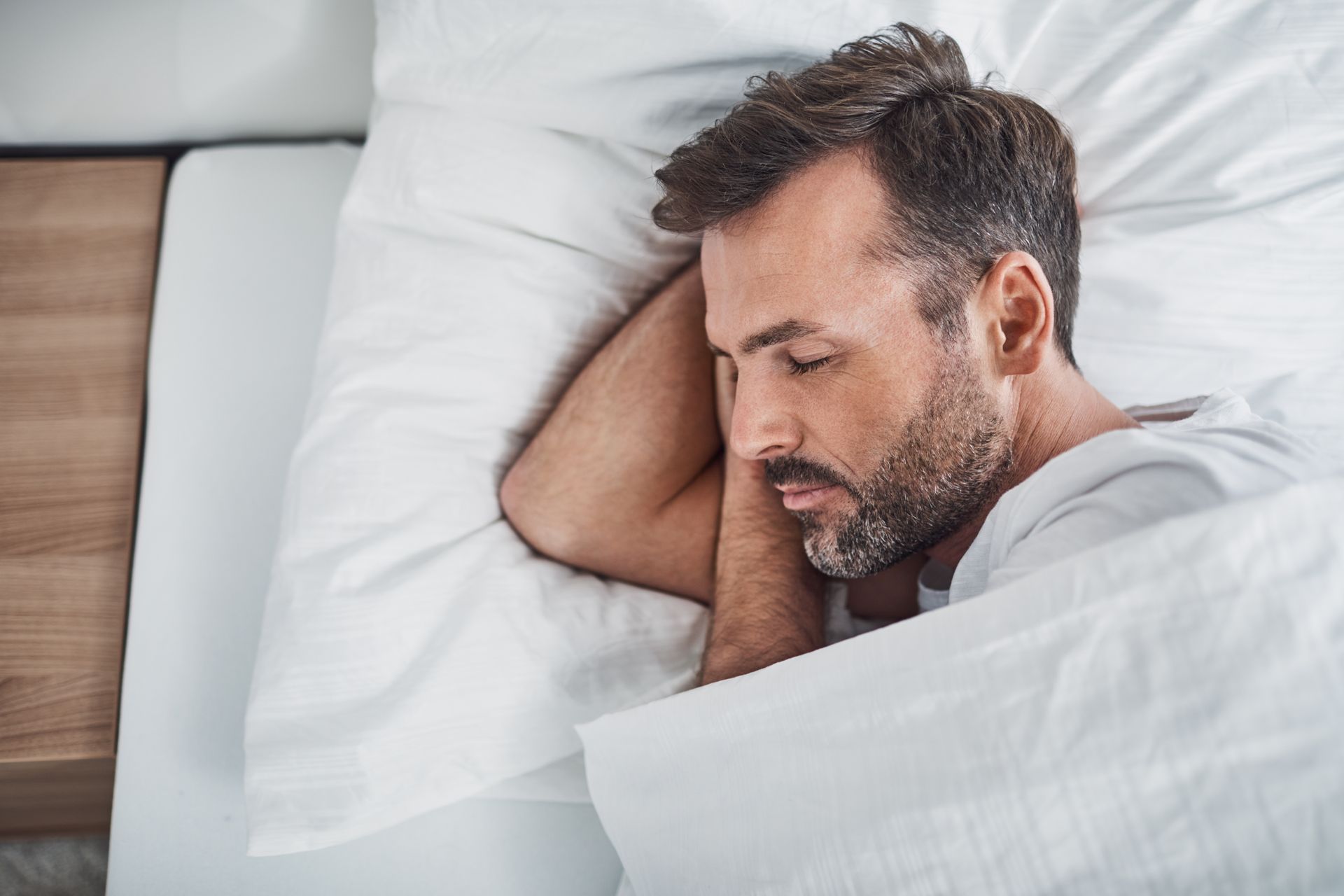 Man sleeping on white bed with eyes closed, resting on his side under white covers