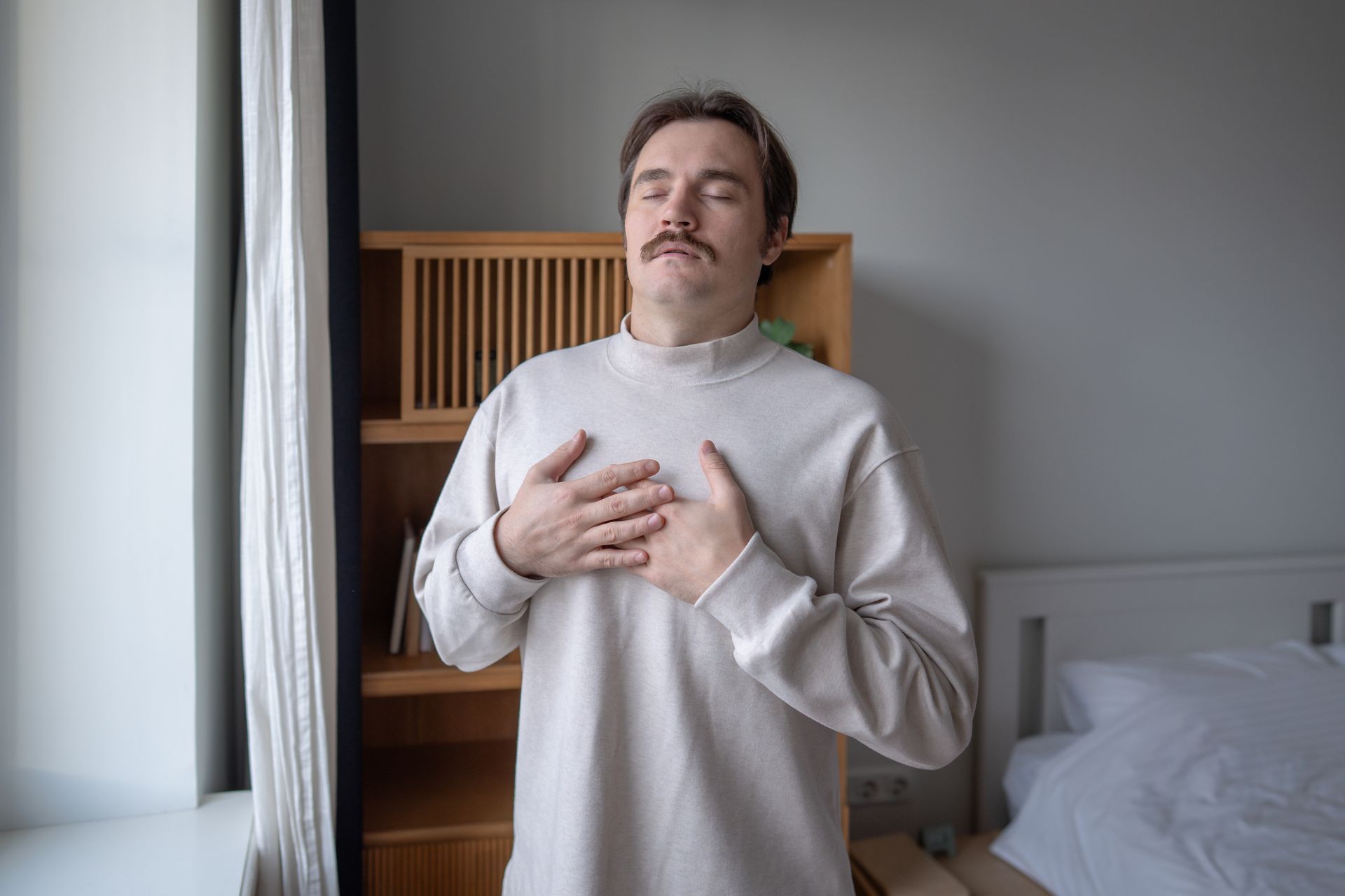 Person standing indoors with hands on chest beside a bed and window in a softly lit bedroom