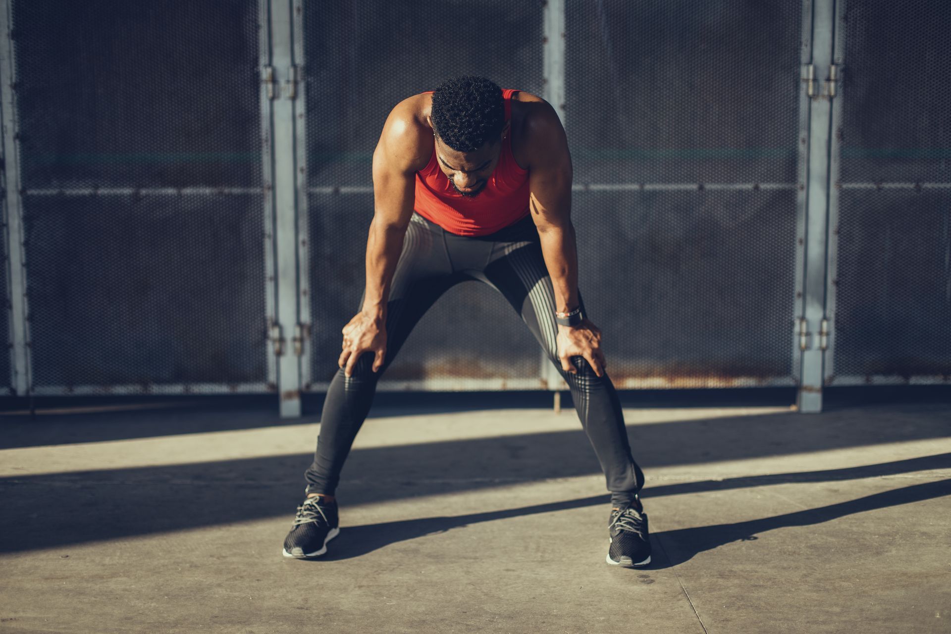 Athlete in red top bent over catching breath after workout on a concrete floor in front of a dark wall