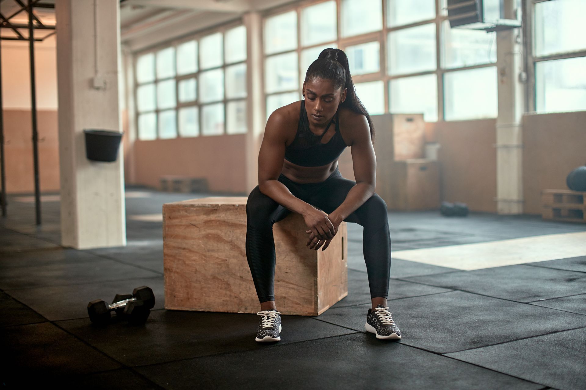 Athlete resting on a wooden box in a gym, hands on knees, with a dumbbell nearby.
