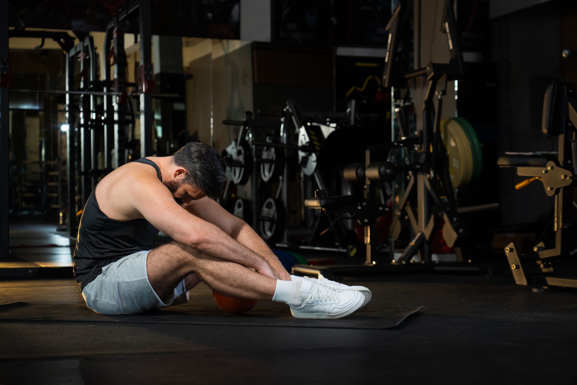Man stretching on a gym mat, seated forward fold in a dim weight room