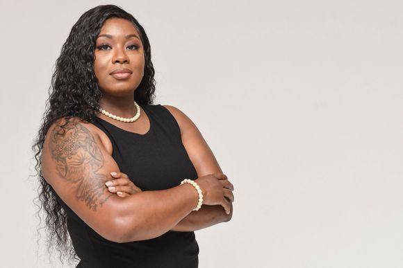Woman with curly hair and tattoos poses with arms crossed against a light gray background