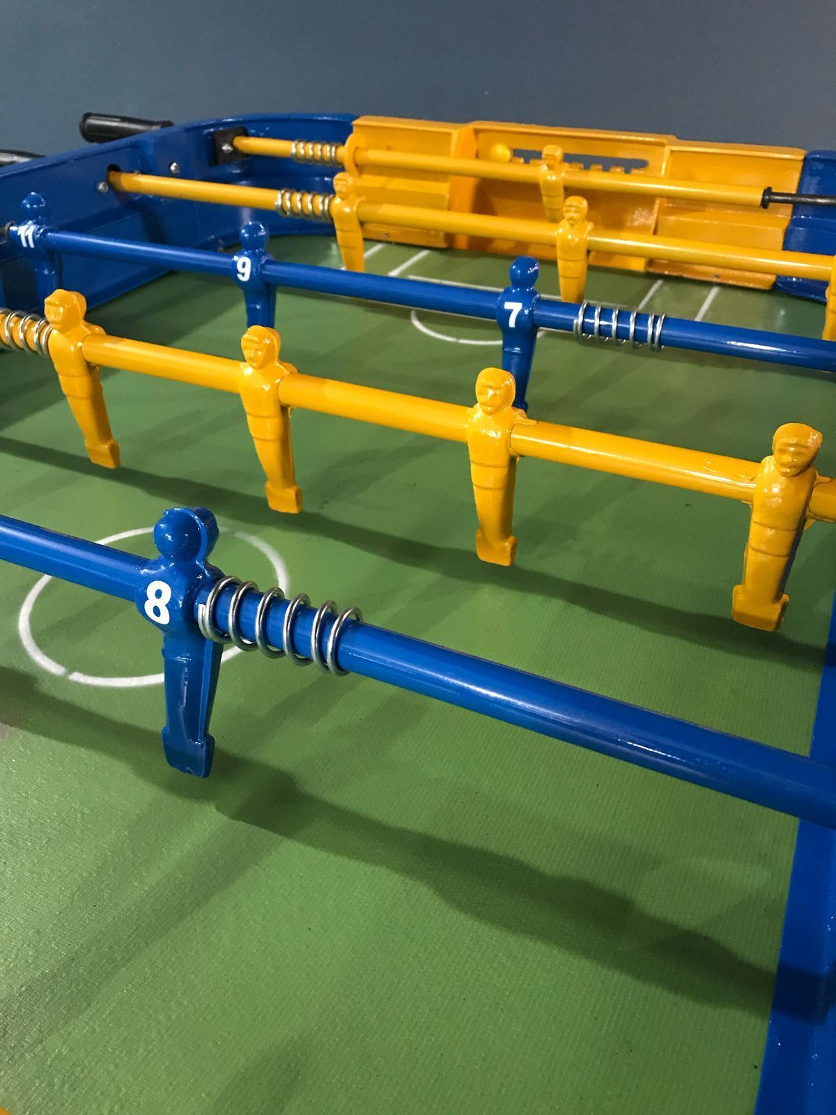 Una mesa de futbolín azul y amarilla con jugadores azules y amarillos numerados en varillas de metal sobre un campo verde.
