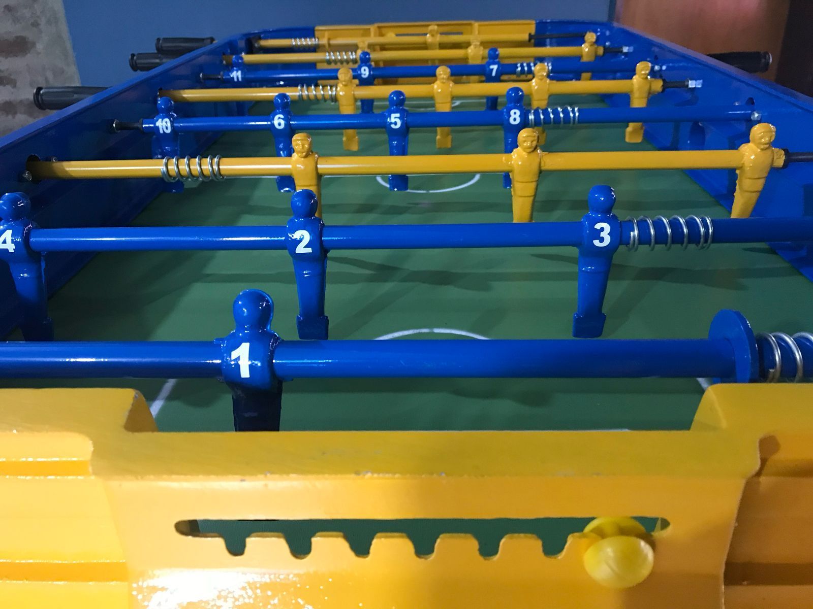 Una mesa de futbolín azul y amarilla con jugadores azules y amarillos numerados en varillas de metal sobre un campo verde.
