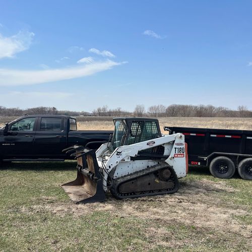 Black pickup truck and white skid steer in a field beside a black trailer under a clear blue sky