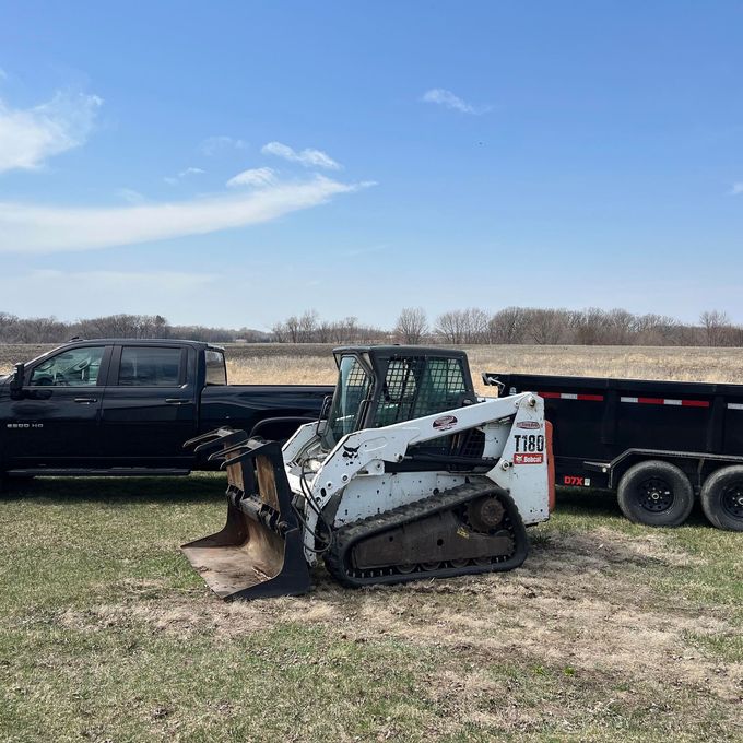 White Bobcat loader beside a black pickup truck and trailer in a grassy field under a blue sky
