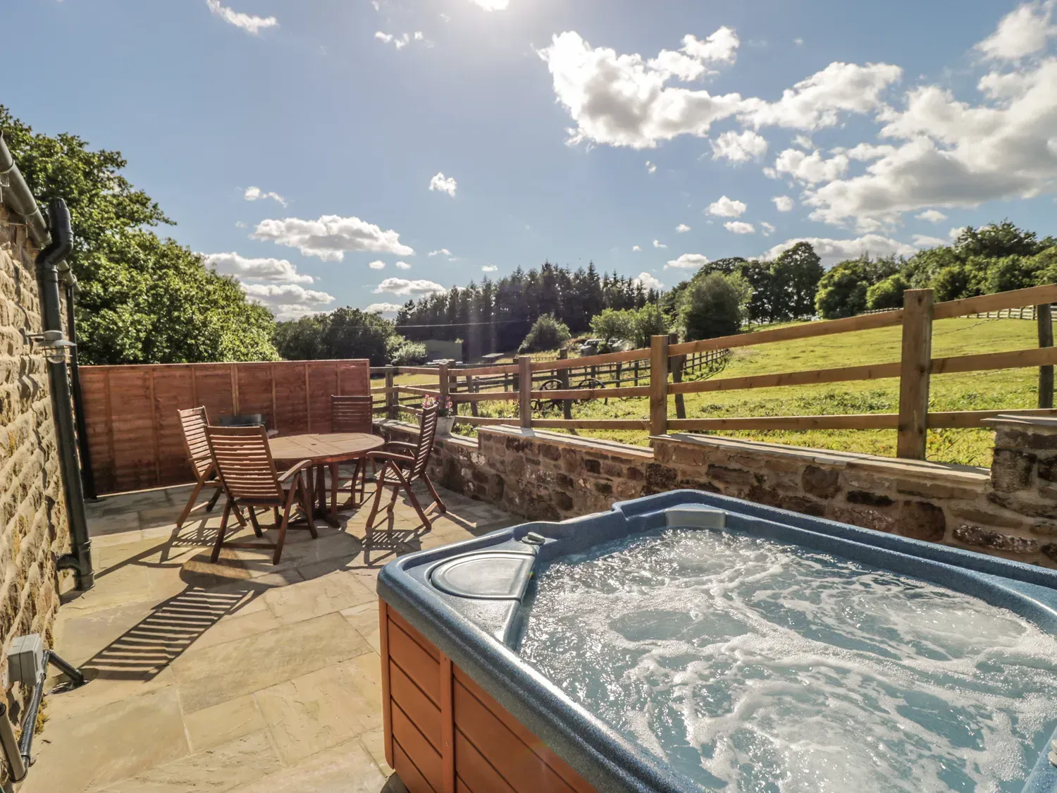 Hot tub on patio overlooking a field with table and chairs. Sunny day, blue sky.