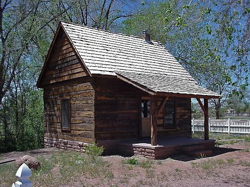 Log cabin with a porch, cedar shake roof, and stone foundation, surrounded by trees and a white fence.