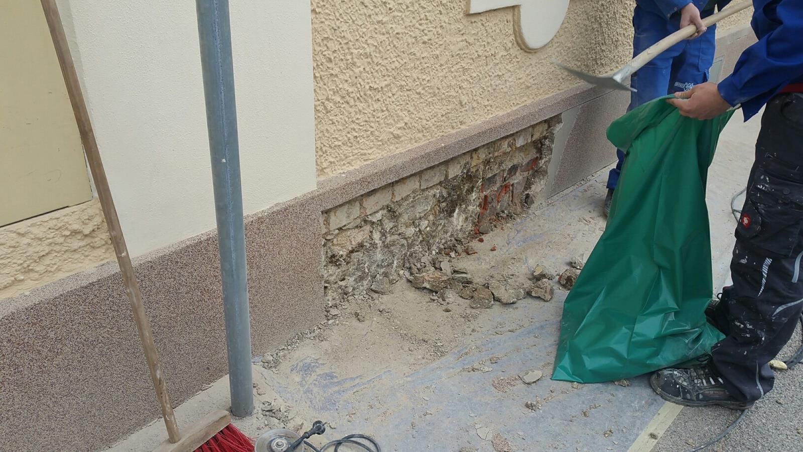 Arbeiter entfernen beschädigte Steinverblendungen vom Sockel einer Gebäudewand und füllen die Trümmer in einen grünen Sack.