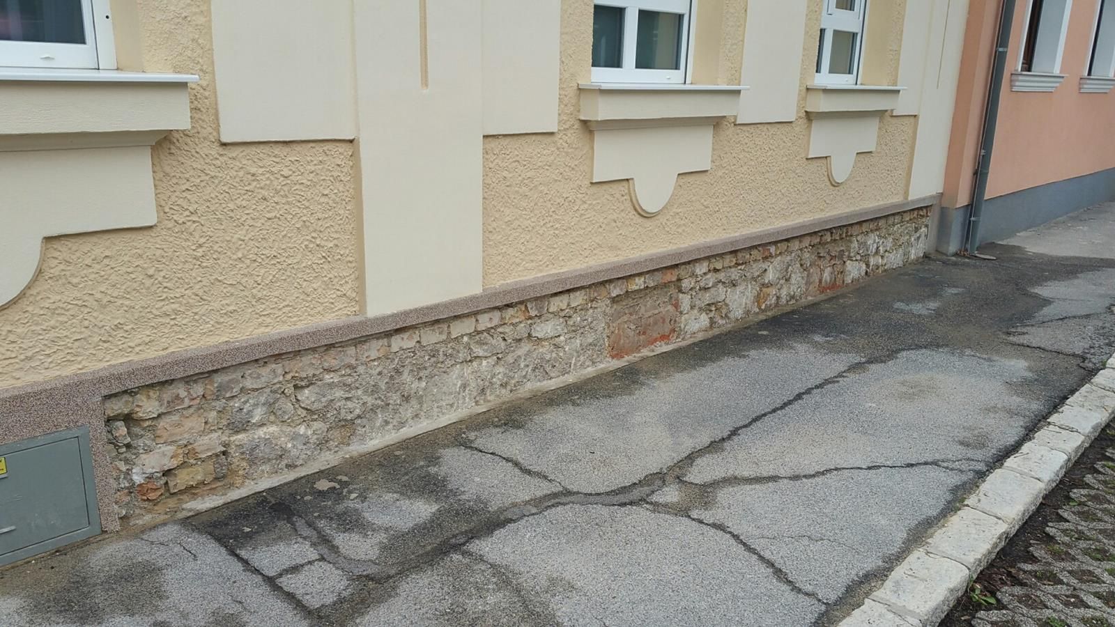Nahaufnahme einer gelben Putzwand über einem verwitterten Steinfundament, die von einem rissigen Asphaltweg begrenzt wird.