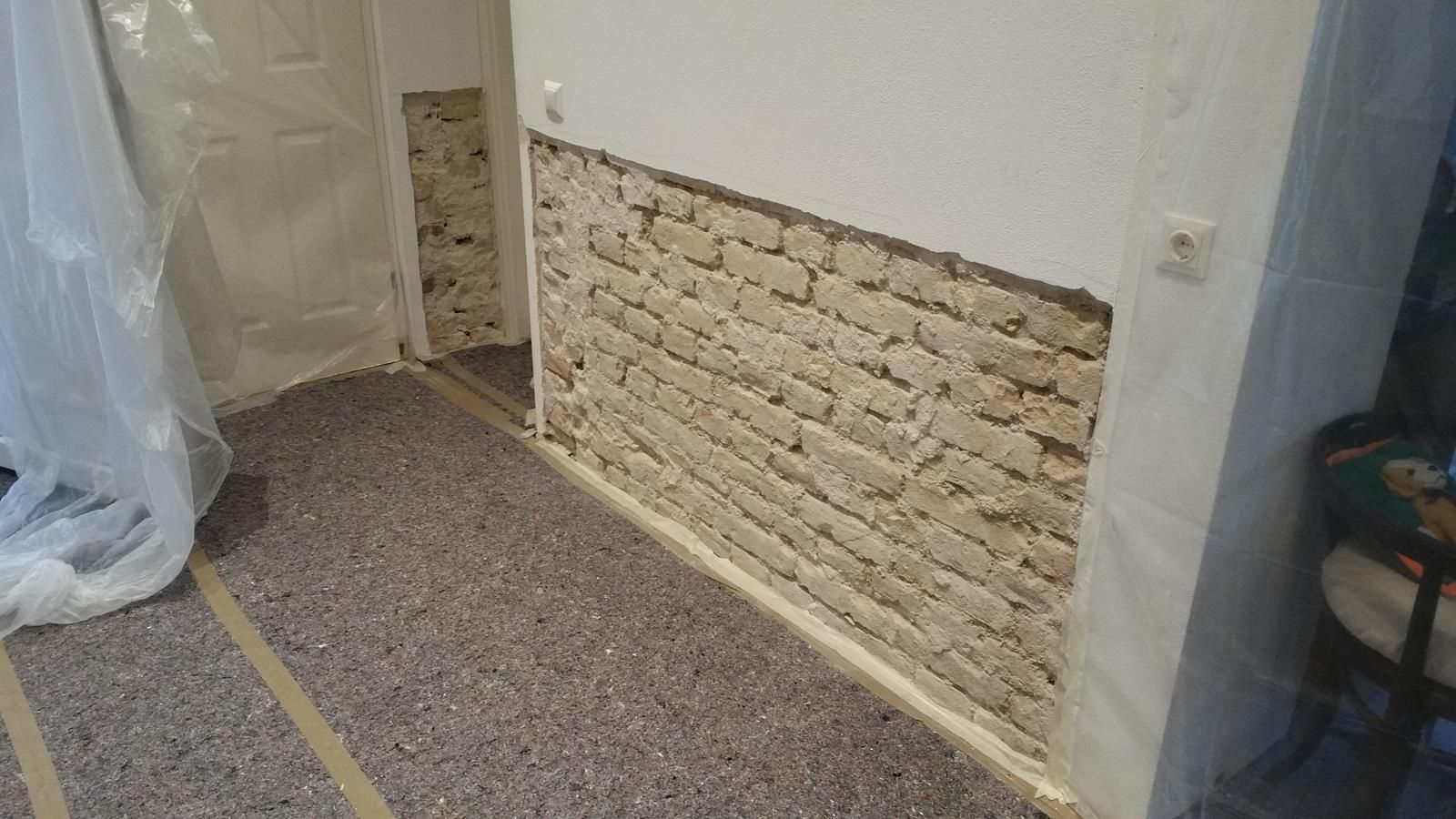 In einem Haus wird ein hellfarbiger Ziegelsteinabschnitt freigelegt, der gerade repariert wird. In der Nähe sind Bodenschutzbänder und eine Tür zu sehen.
