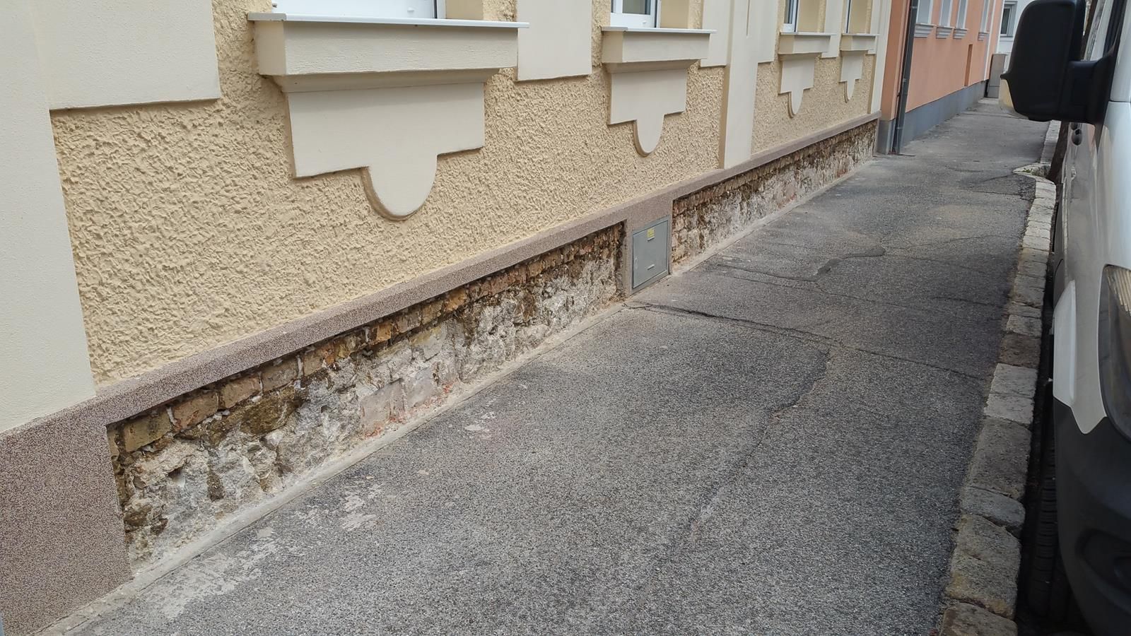 Eine hellbraune Hauswand mit beschädigtem, freiliegendem Ziegelsockel neben einem grauen Asphalt-Gehweg.