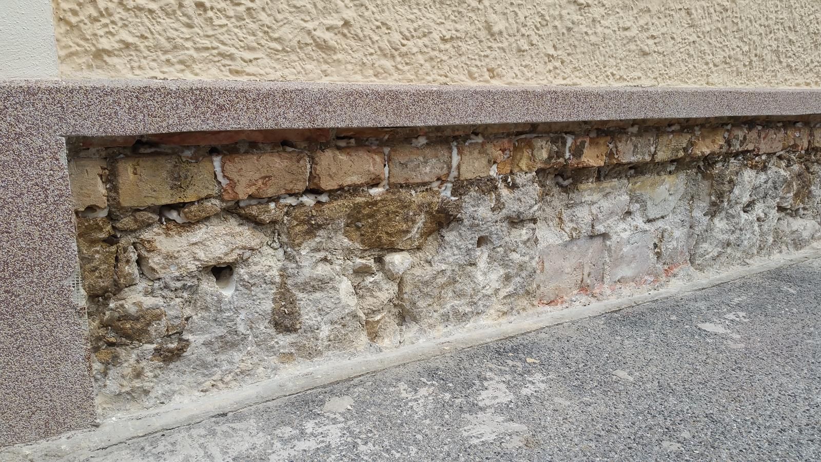 Beschädigtes Beton- und Ziegelfundament einer Gebäudewand, dessen bröckelnde Struktur über einem grauen Gehweg sichtbar ist.