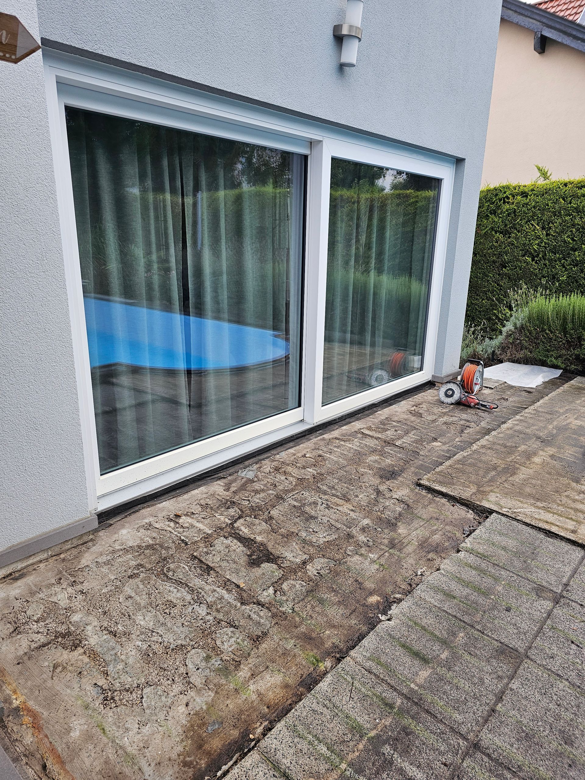 Schiebetüren aus Glas führen auf eine Terrasse mit freiliegendem Betonsockel und angrenzender Hecke und Swimmingpool.