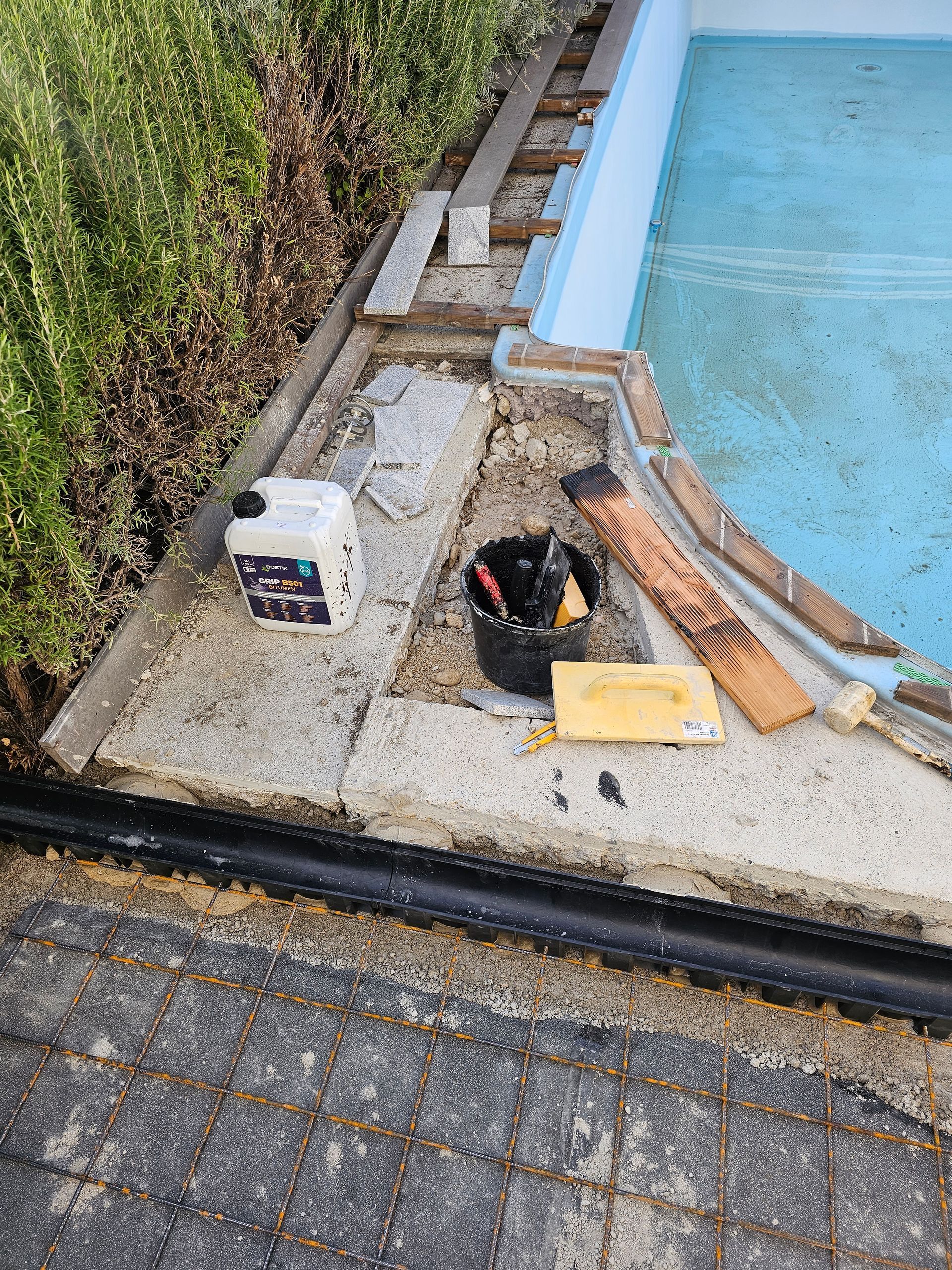 Werkzeuge, Reinigungsmittel und Bauschutt auf einer Betonpoolterrasse in der Nähe eines blau gestrichenen Poolrandes und steinerner Terrassenplatten.