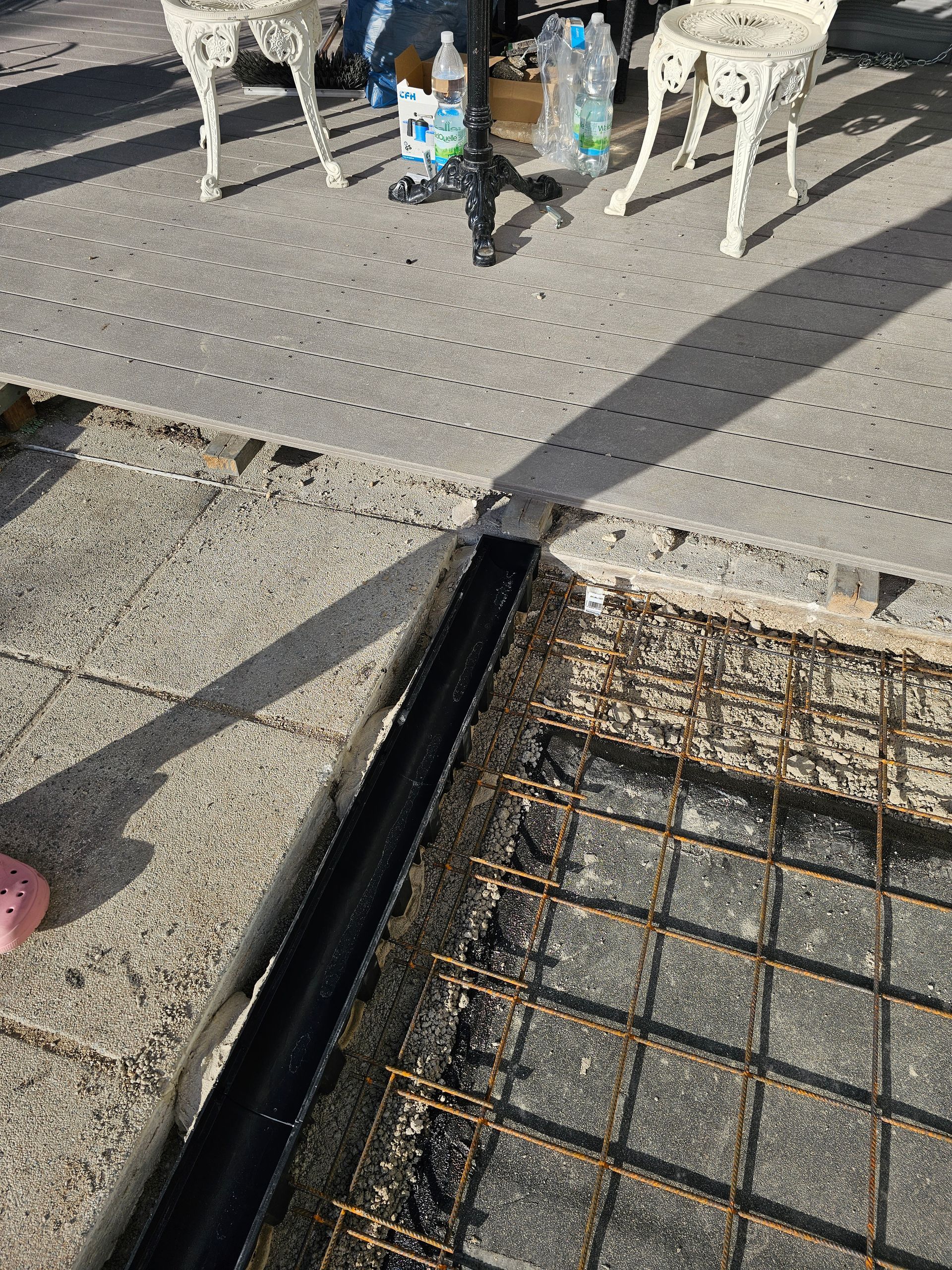 Zwischen einer Betonplatte und einer Baustelle mit freiliegendem Drahtgitter verläuft ein schwarzer Metallablauf.