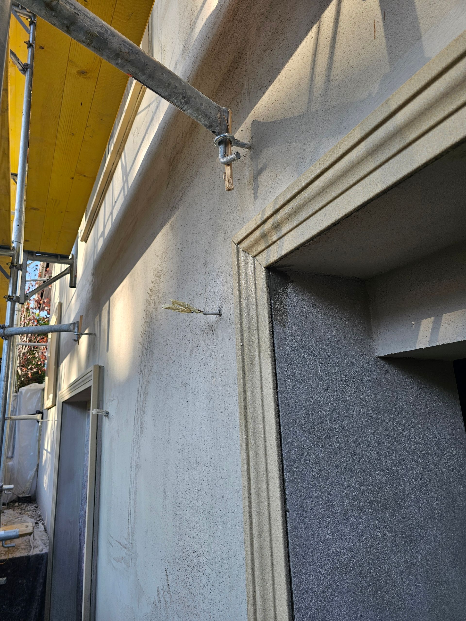 An einer strukturierten Außenwand neben einem im Bau befindlichen Bogeneingang ist ein Gerüst befestigt.