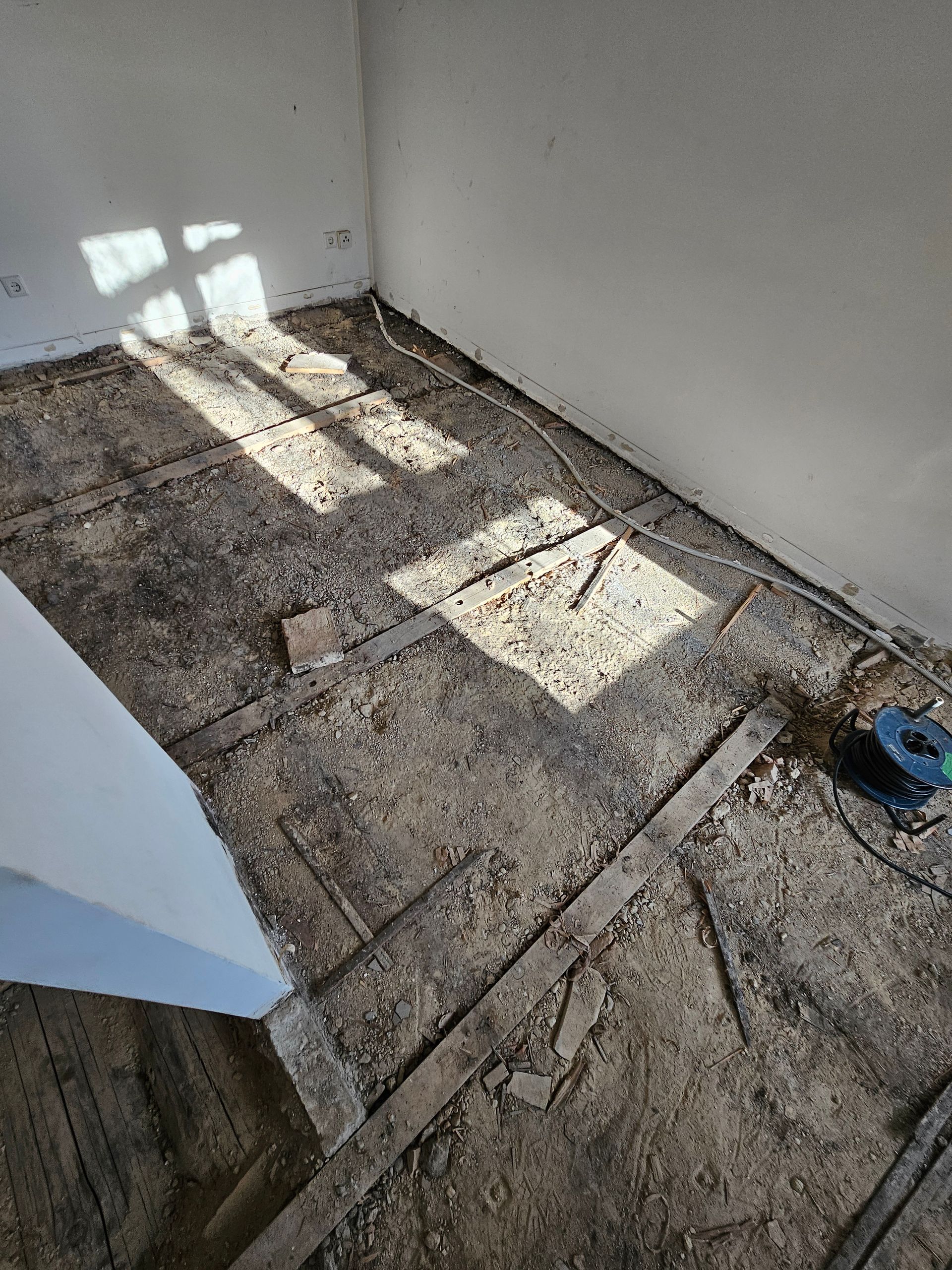 Ein unfertiger Raum mit einem Unterboden aus freiliegenden Holzbalken, der mit Schutt und Staub bedeckt ist, erleuchtet vom Licht eines Fensters.