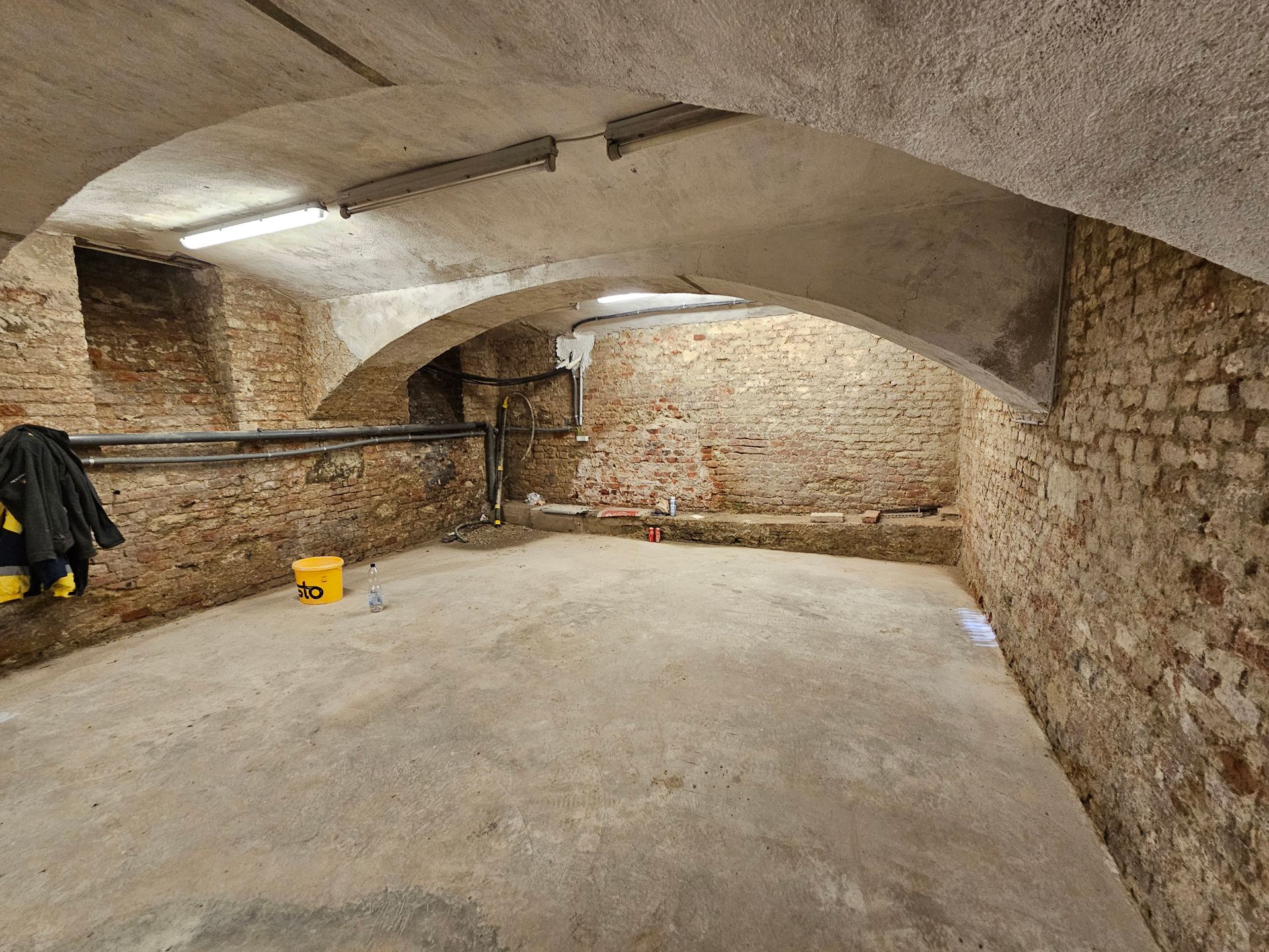 Ein leerer, von Steinmauern umgebener Kellerraum mit Gewölbedecke, Betonboden und freiliegenden Rohren an der Seitenwand.