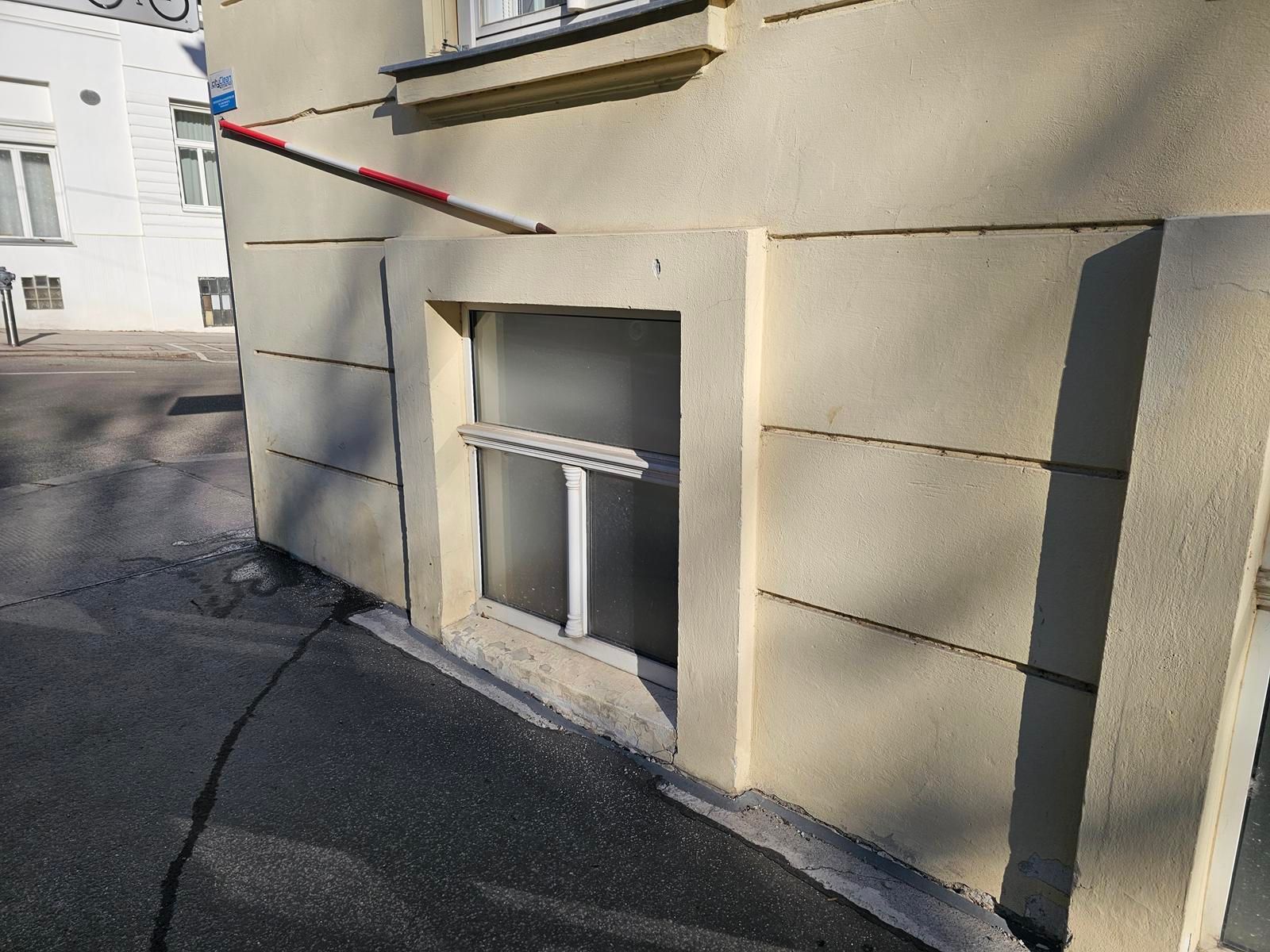 Ein rot-weißer Absperrpfosten ragt von der beigen, strukturierten Wand eines Gebäudes über einem ebenerdigen, mattierten Fenster hervor.