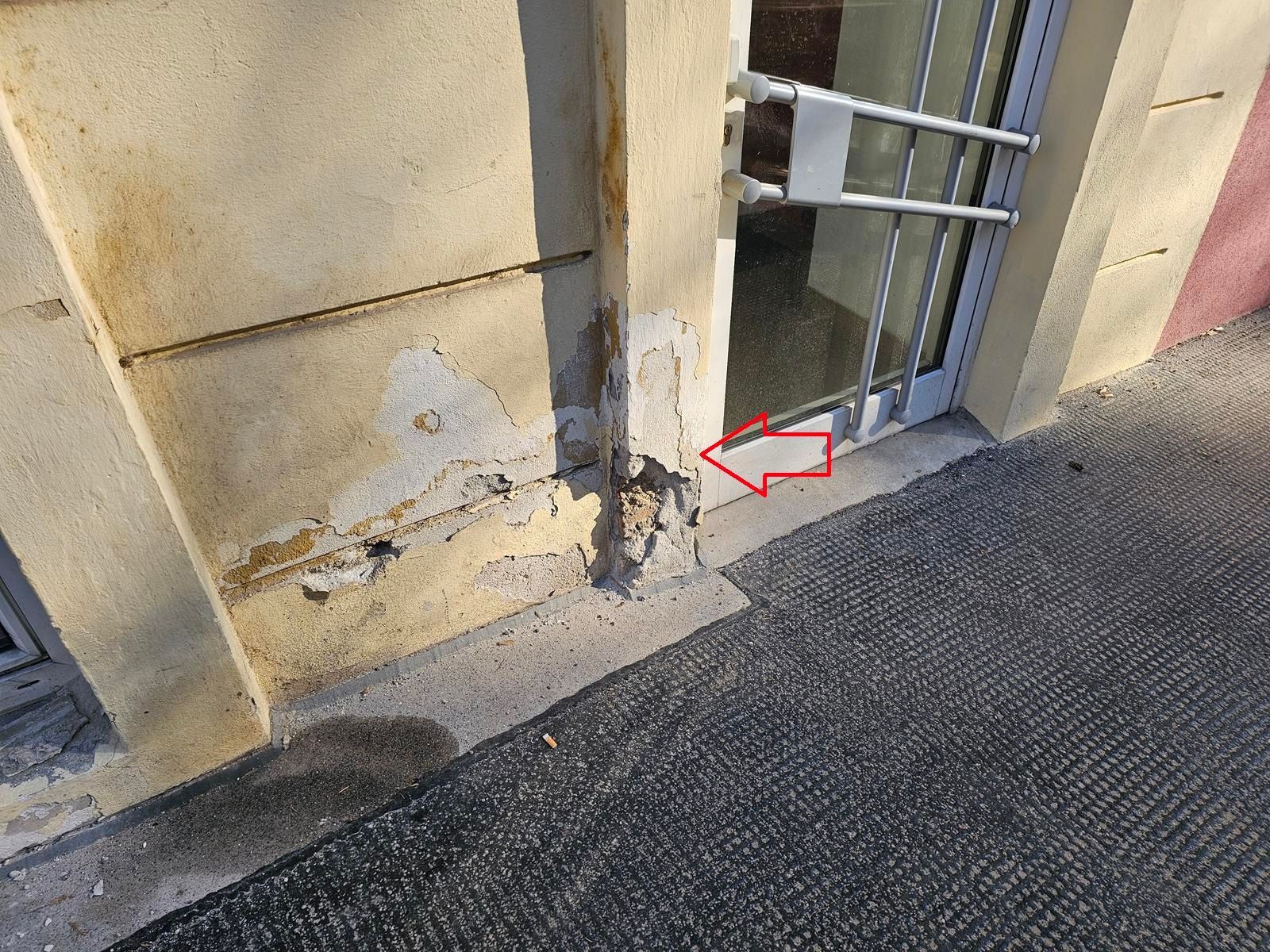 Ein roter Pfeil weist auf einen beschädigten, bröckelnden Abschnitt einer hellen Außenwand in der Nähe eines Türrahmens aus Metall und des Gehwegs.