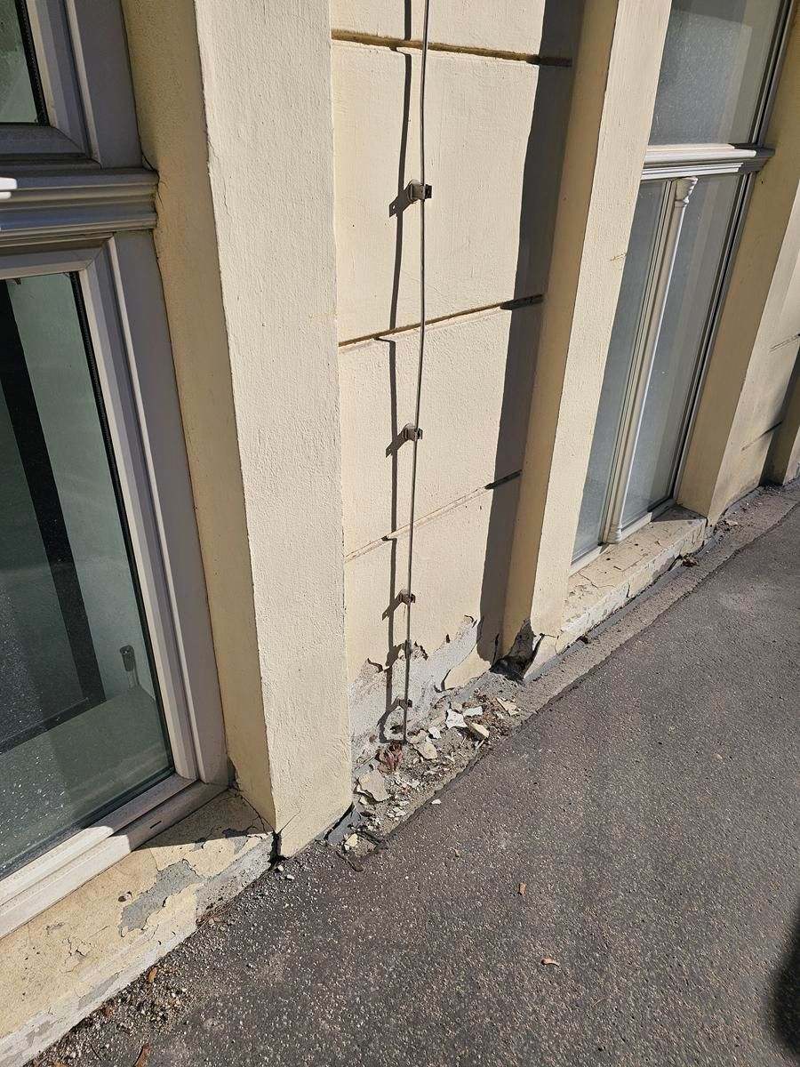 Ein senkrechter Draht oder ein Kabel, das an einer beigen Außenwand befestigt ist, wobei der Beton am unteren Ende in Bodennähe brüchig ist.
