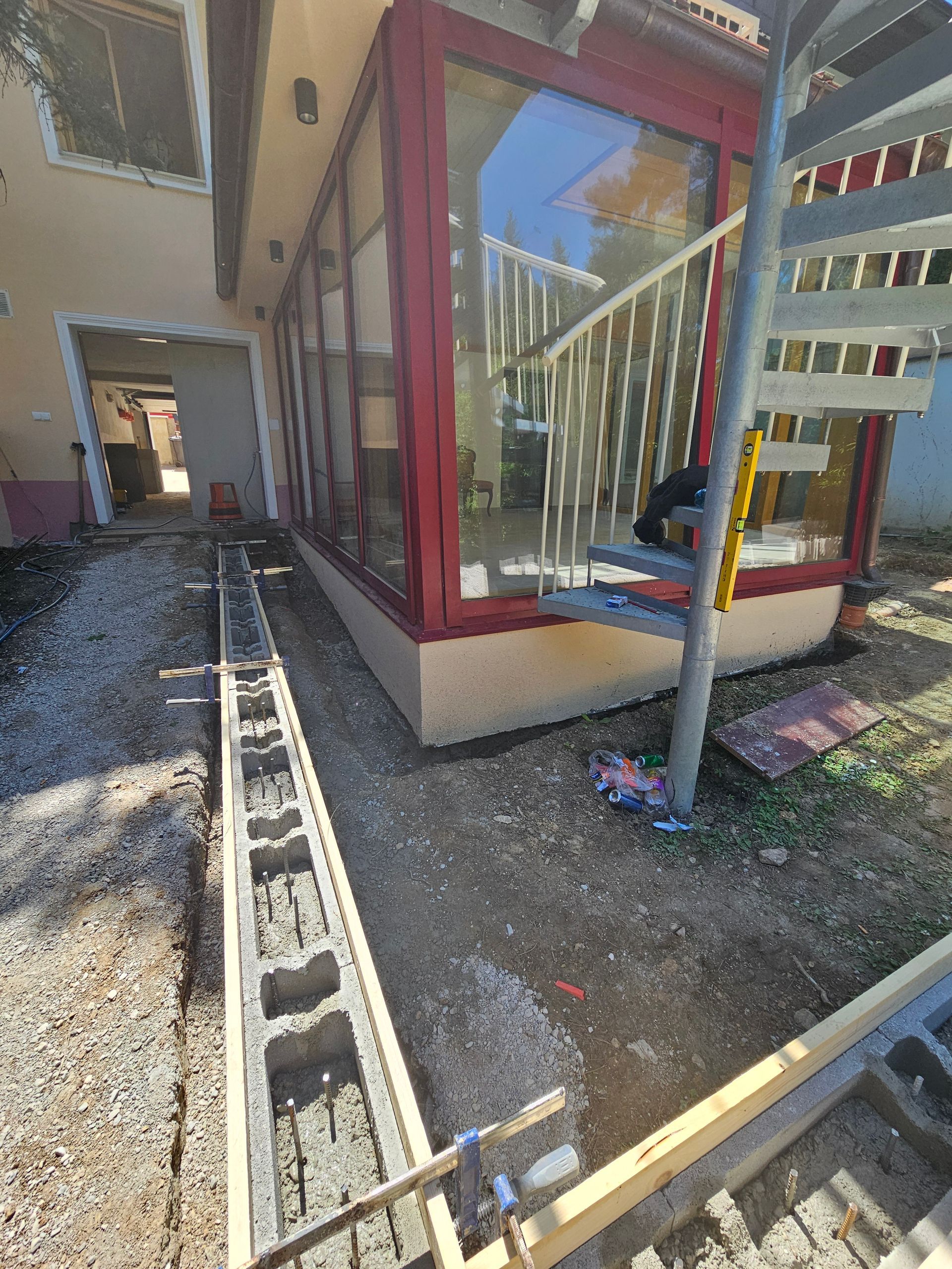 Eine im Bau befindliche Betonsteinmauer neben einem Haus mit einem rot umrahmten Wintergarten und einer Wendeltreppe.