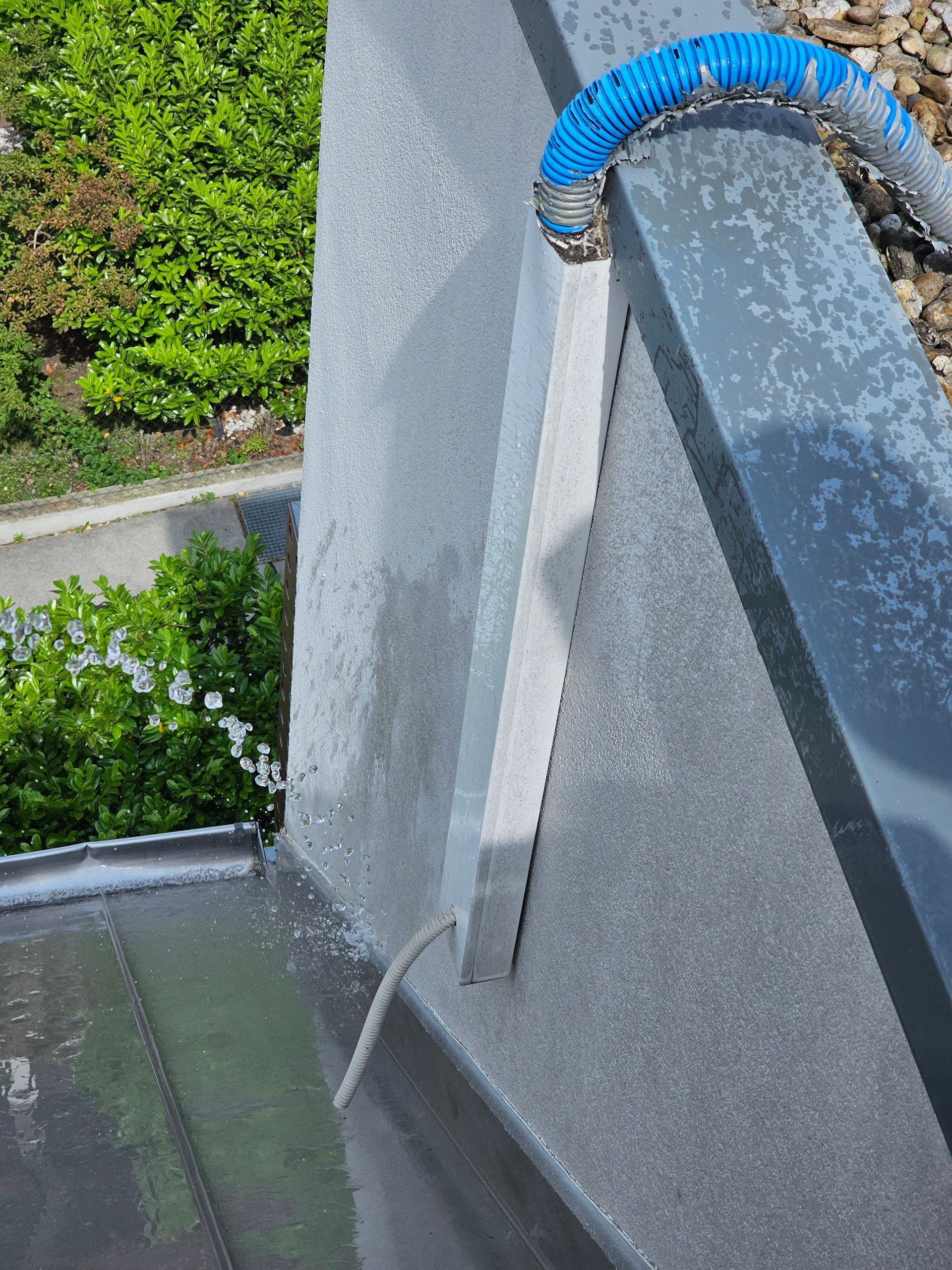 Ein blauer Schlauch verläuft entlang einer grauen Betonwand und mündet durch einen Metallkanal auf ein darunter liegendes Dach.