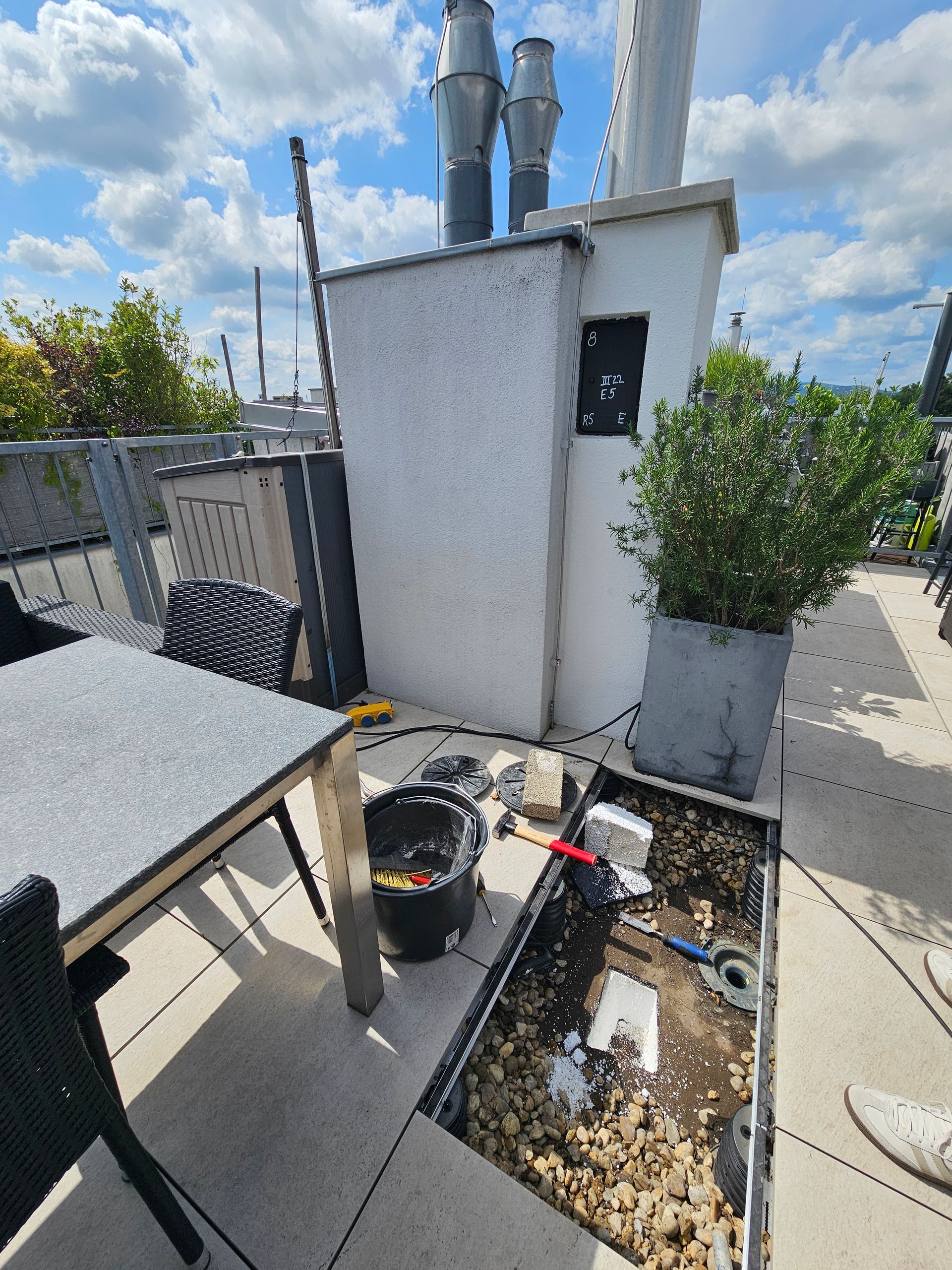 Eine Dachterrasse mit Tisch und Stühlen, über der sich ein offener, mit Erde gefüllter Abflussgraben mit Werkzeugen und Unrat befindet.