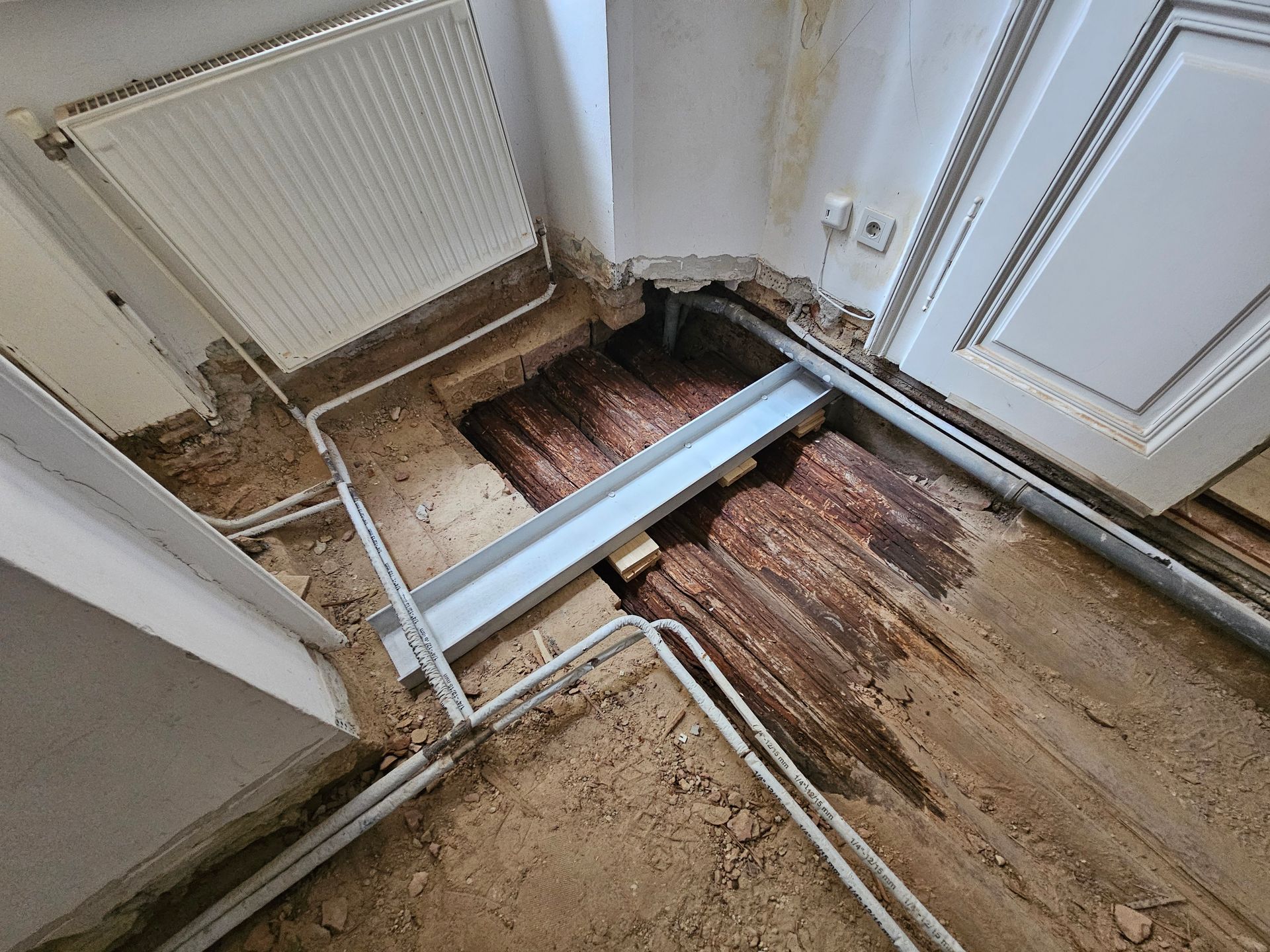 Bei einer Hausrenovierung wurde ein beschädigter Holzboden freigelegt, auf dem in der Nähe eines Heizkörpers verlegte Metallrohre und ein Stützbalken zu sehen sind.