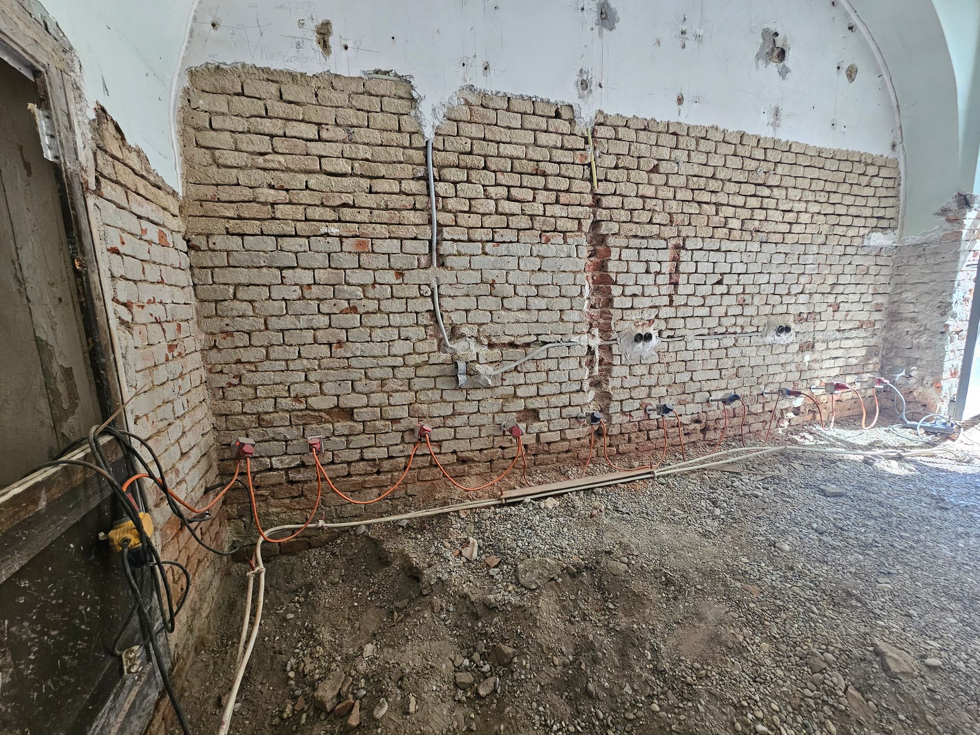 Eine Baustelle im Inneren eines Raumes mit Kiesboden, an der eine freiliegende Ziegelwand mit elektrischen Leitungen zu sehen ist.