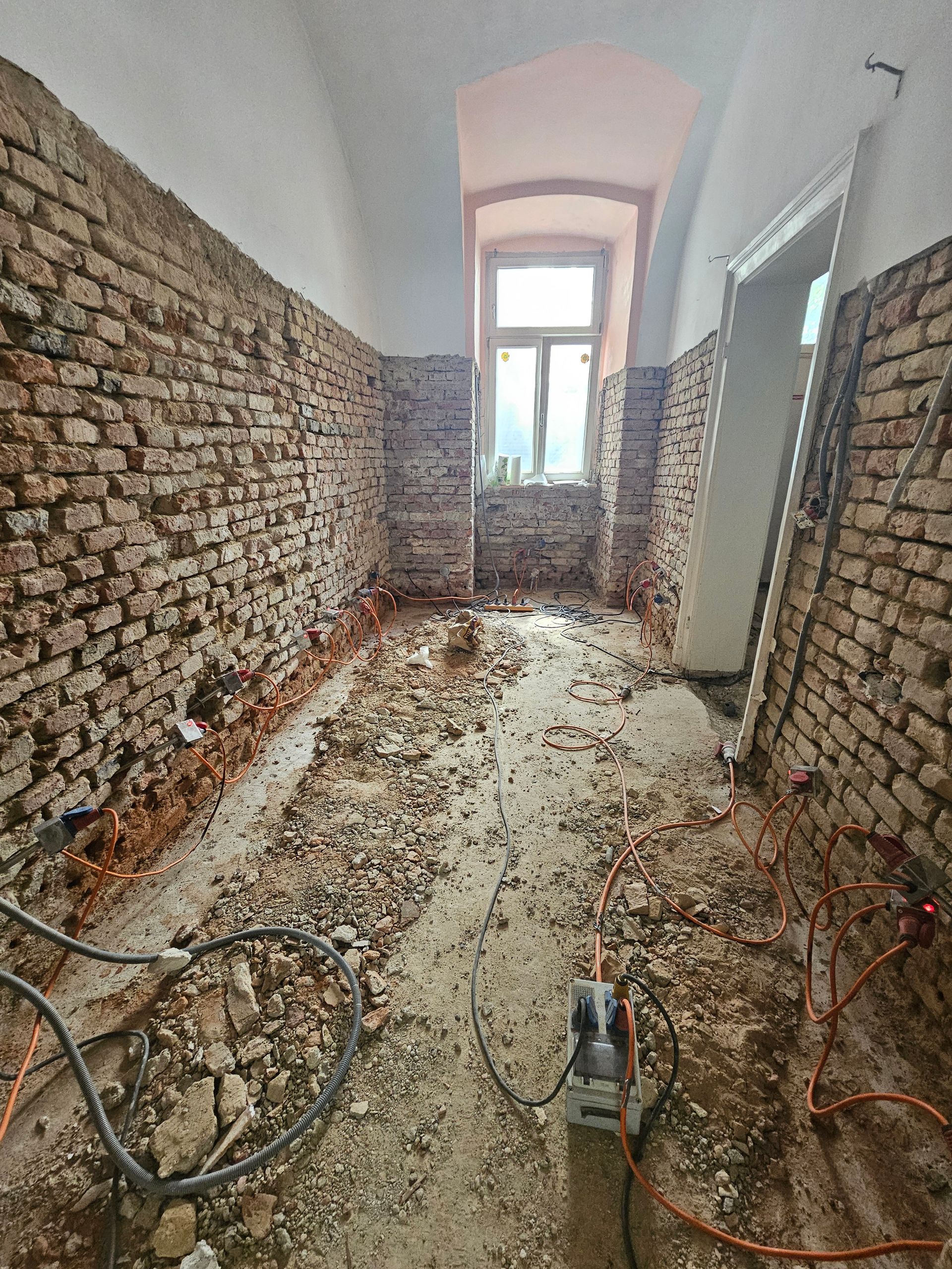 Ein schmaler, im Bau befindlicher Flur mit freiliegenden Ziegelwänden, Bauschutt auf dem Boden und einem Fenster am anderen Ende.