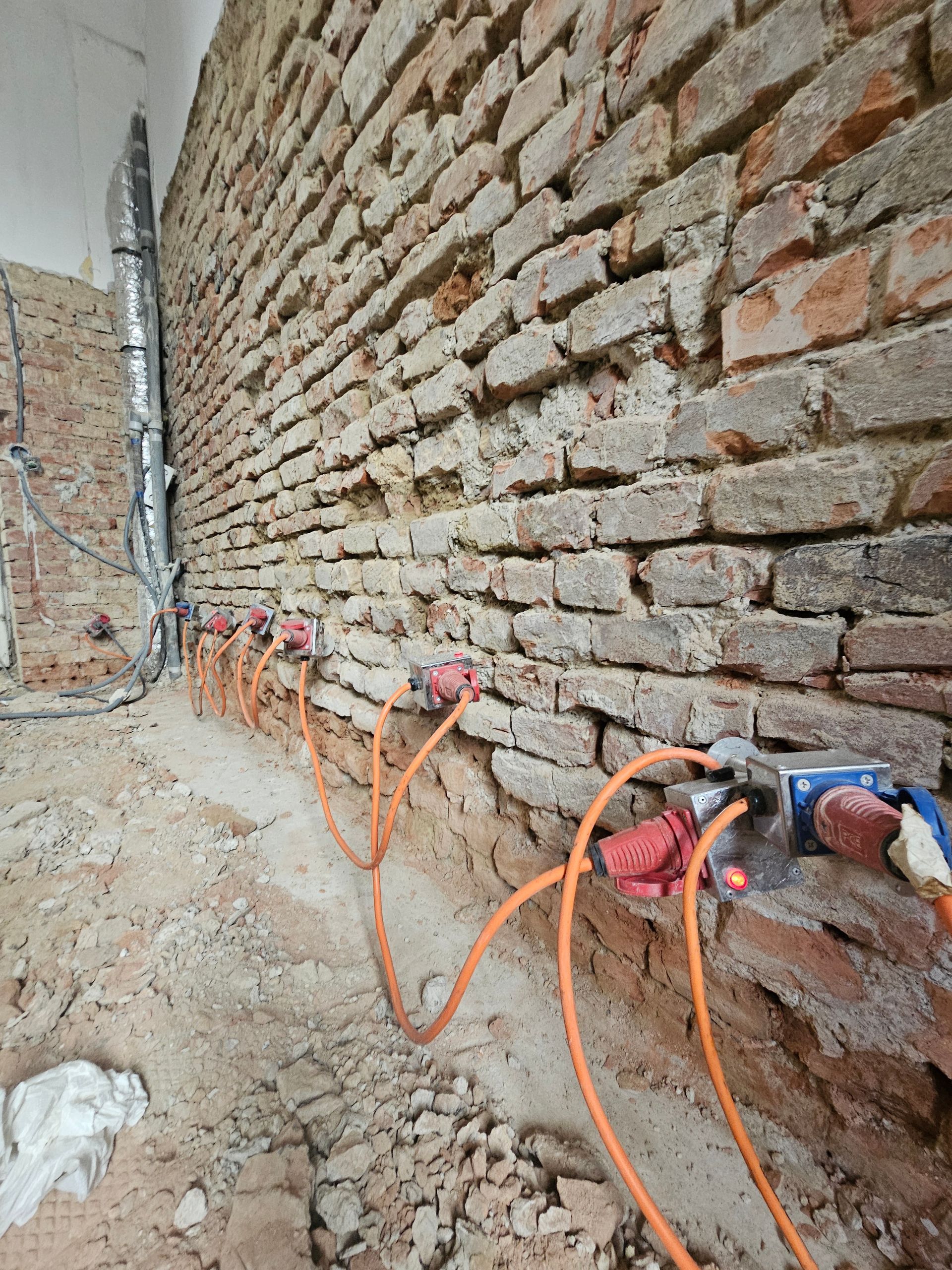 Eine im Bau befindliche, unverputzte Ziegelmauer, an deren Sockel mehrere orangefarbene Elektrokabel mit Verteilerkästen verbunden sind.