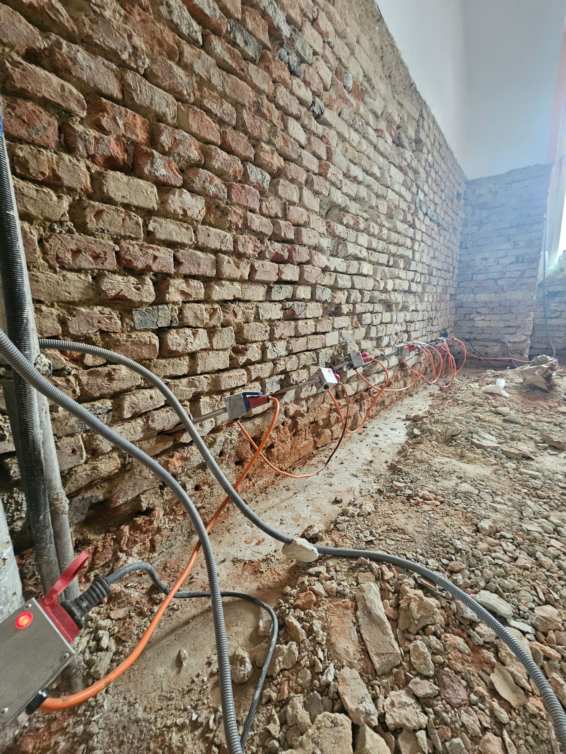 Blick auf eine freigelegte, alte Backsteinmauer während eines Bauprojekts, an deren Sockel elektrische Leitungen verlaufen.