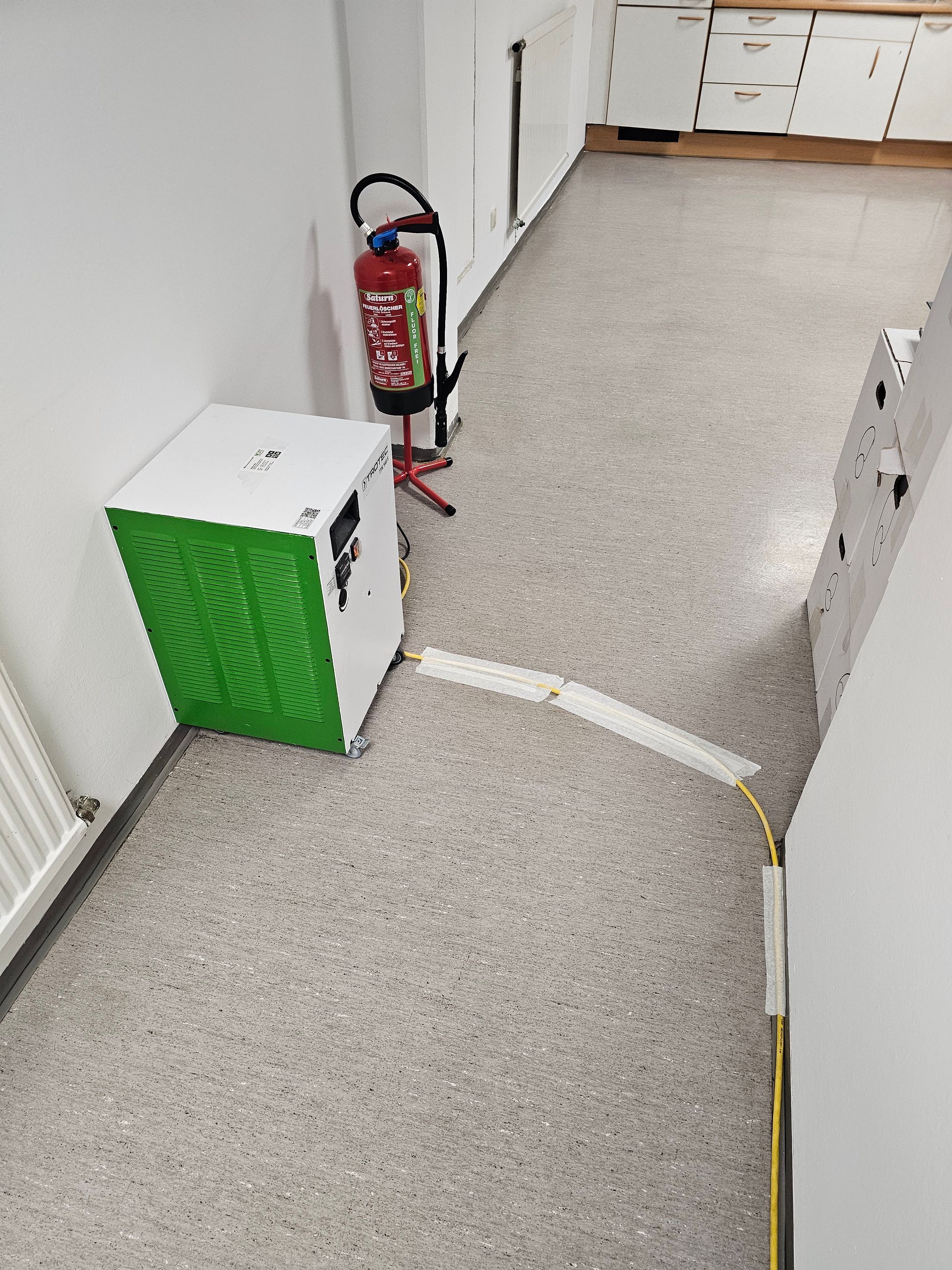 In einem Raum mit einer Kabelabdeckung am Boden steht neben einem Feuerlöscher ein weißer Luftentfeuchter mit grünem Lüftungsschlitz.