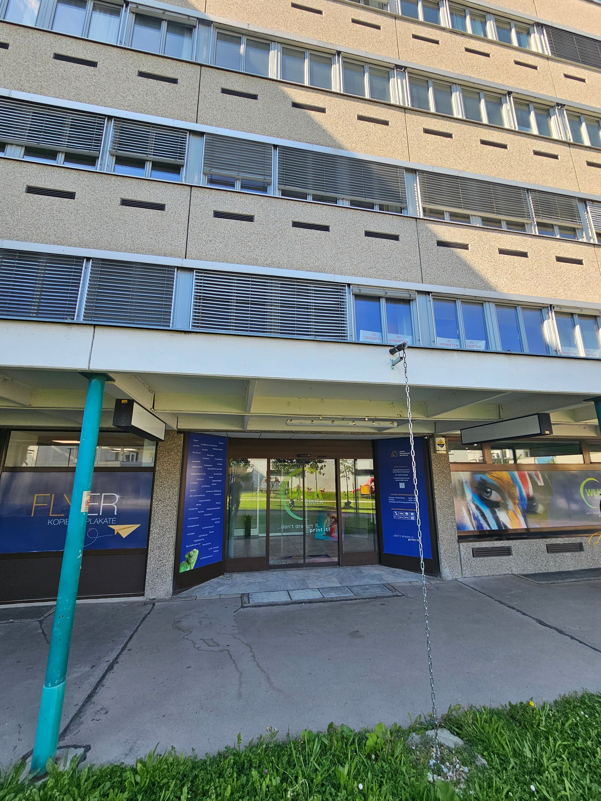 Eine Aufnahme aus Augenhöhe zeigt den Eingang eines Gebäudes mit blau umrandeten Glastüren, großen Fenstern und einer auskragenden Betonfassade.